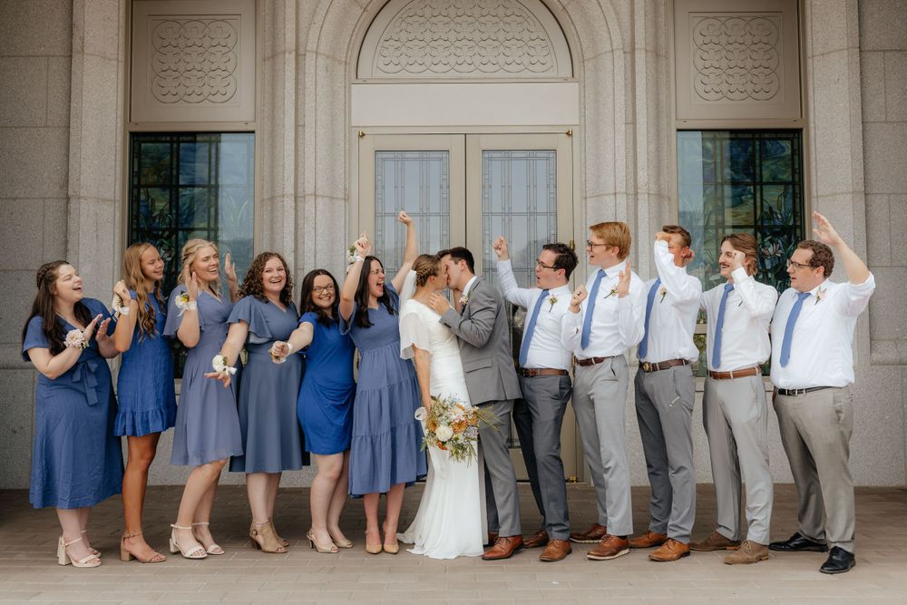 Bride and groom kiss while bridesmaids in blue dresses and groomsmen in gray suits cheer around them.
