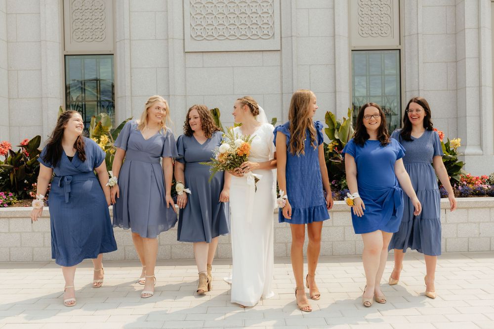 Bride with orange and white bouquet walks with six bridesmaids in blue dresses outside a temple.
