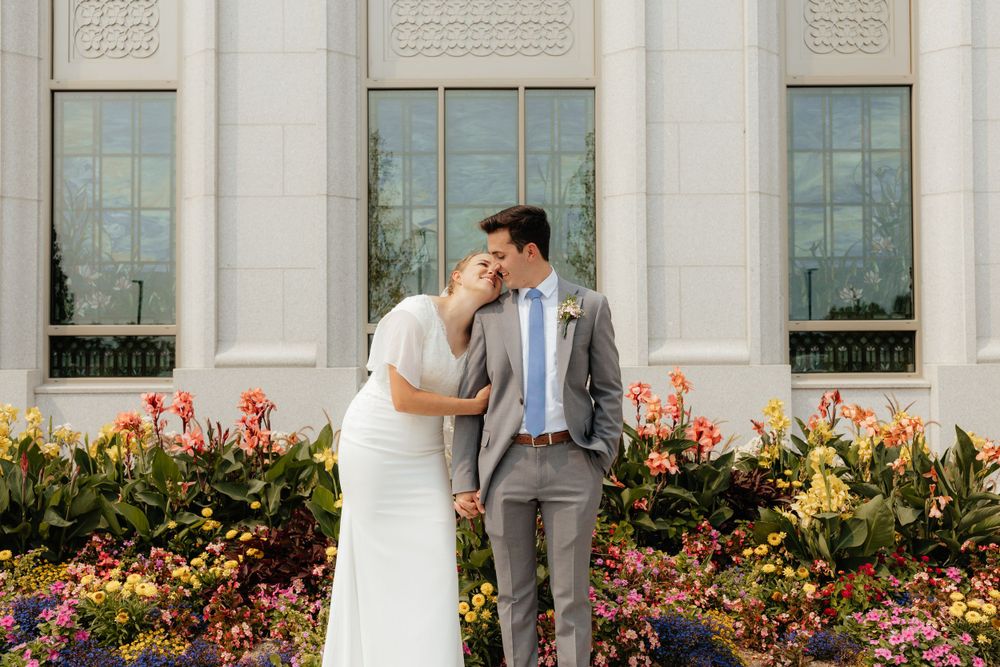 Bride and groom smiling at each other in a garden with colorful flowers outside a white temple.