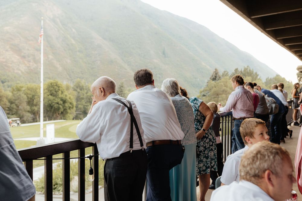 Wedding guests gathered on outdoor balcony overlooking scenic mountain landscape.