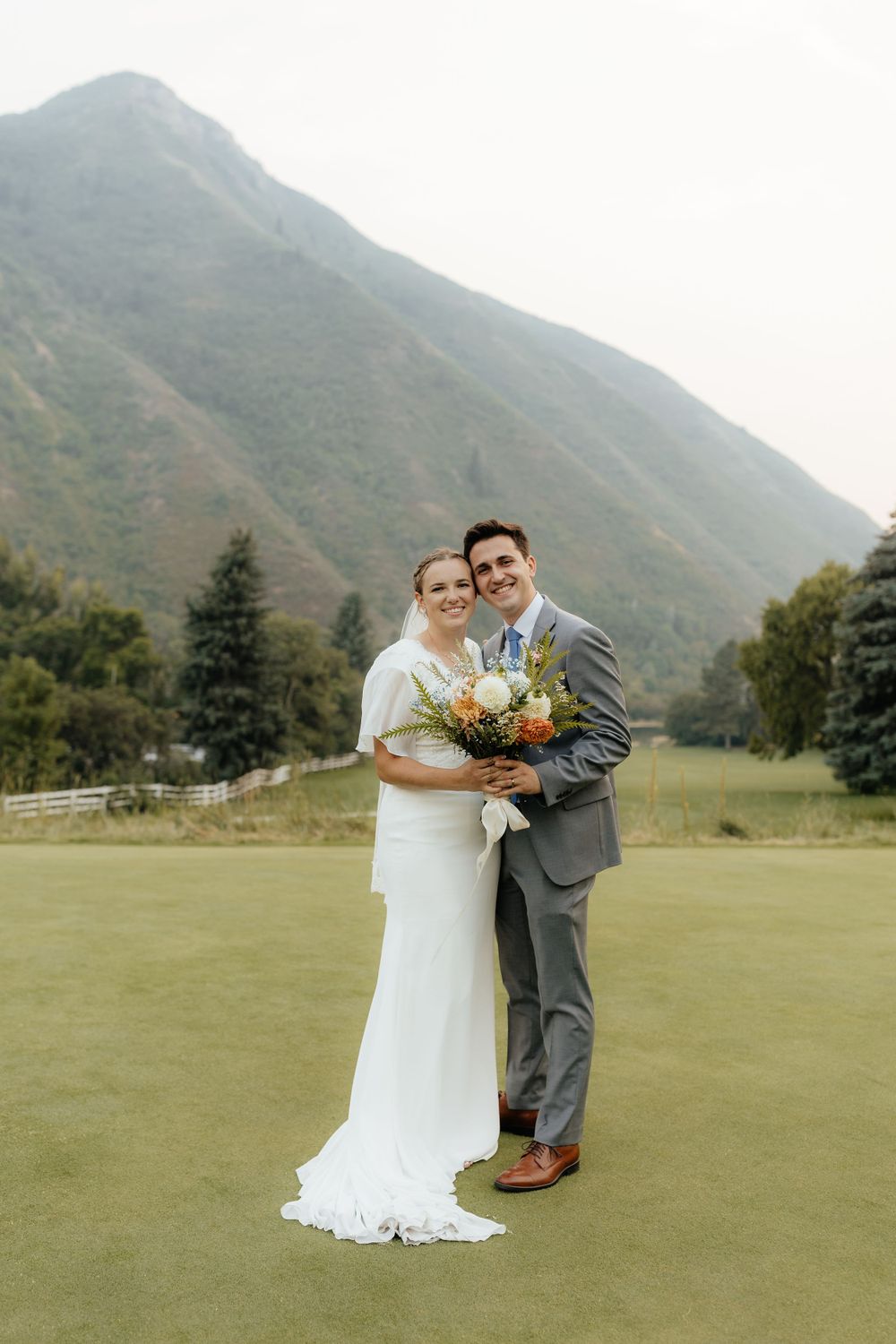 Smiling bride and groom posing on a green golf course with a large mountain backdrop.