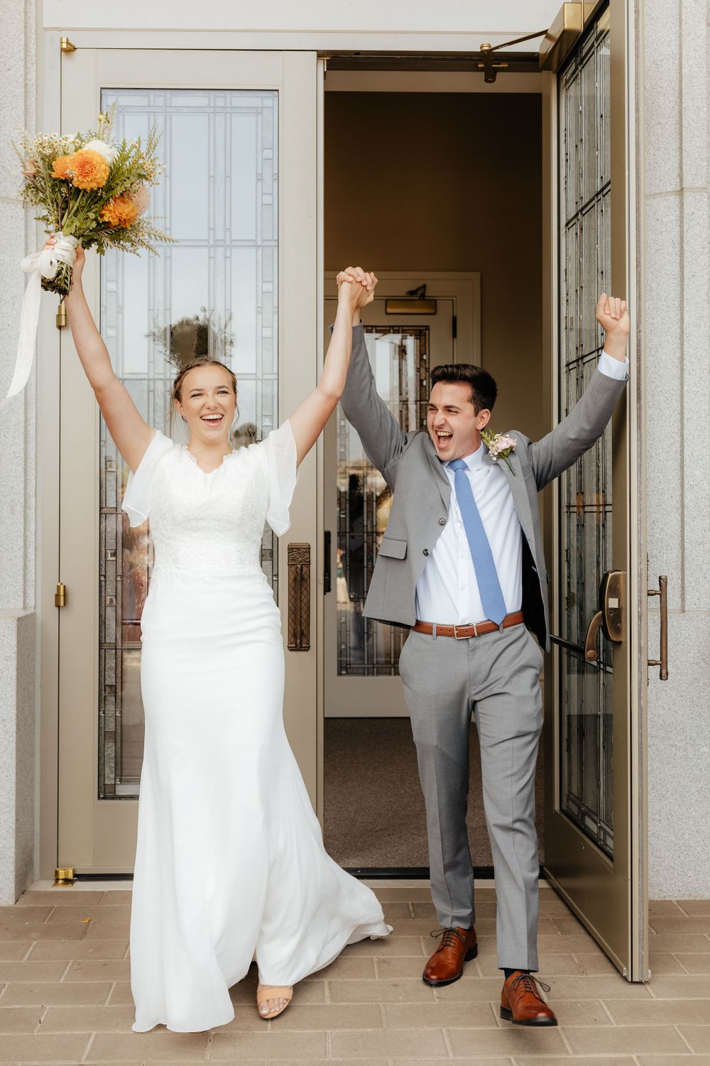 Joyful bride and groom raising arms in celebration exiting wedding venue doors.