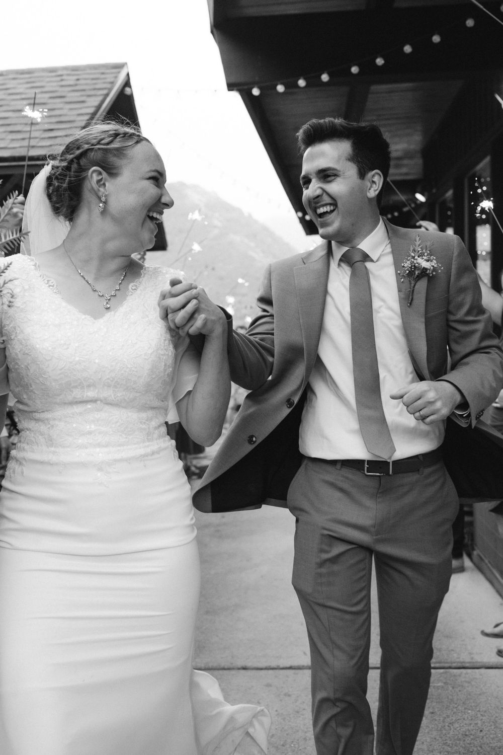 Happy bride and groom laugh together holding hands in black and white outdoor wedding photo.