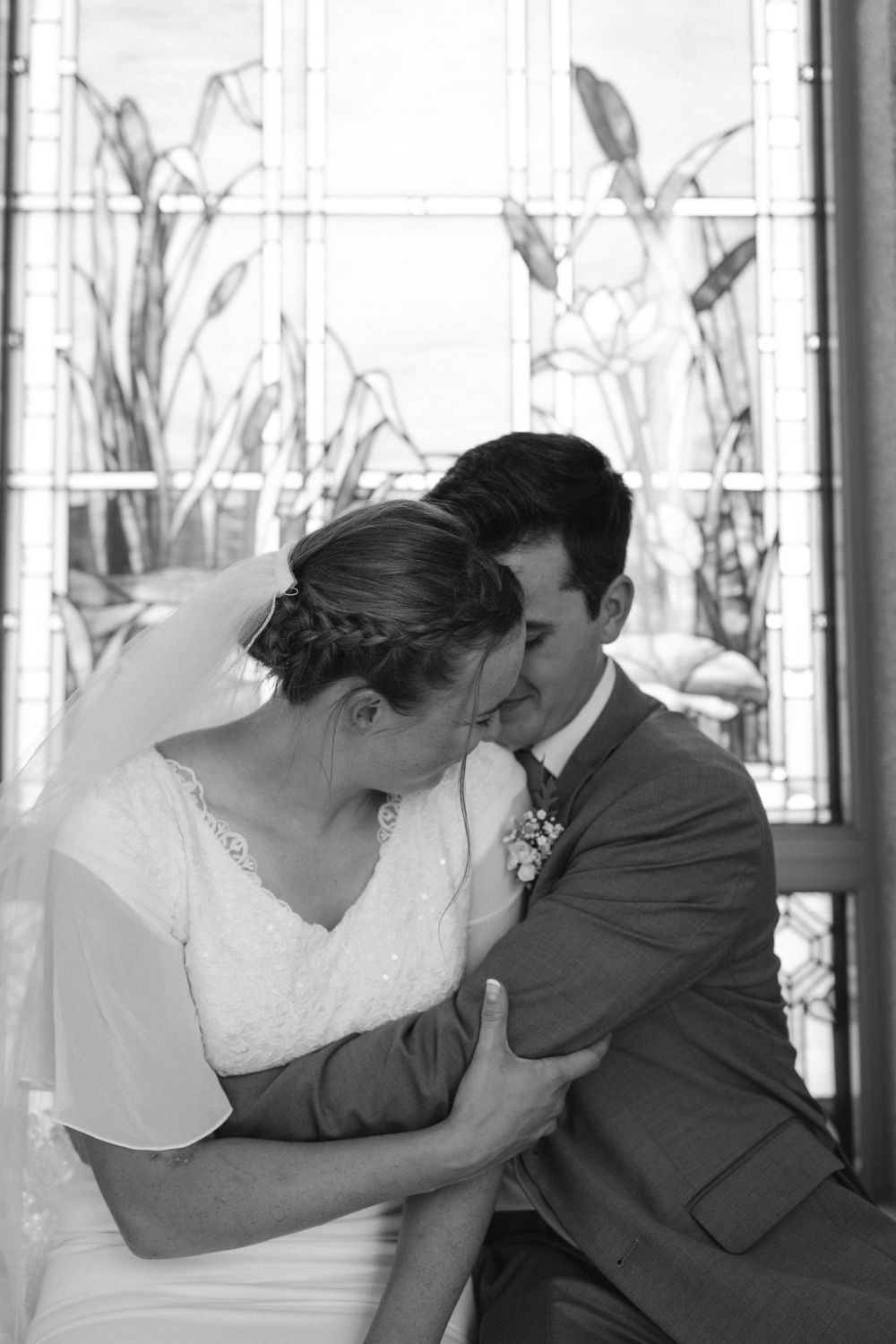 Black and white portrait of bride and groom sharing intimate kiss by stained glass window.
