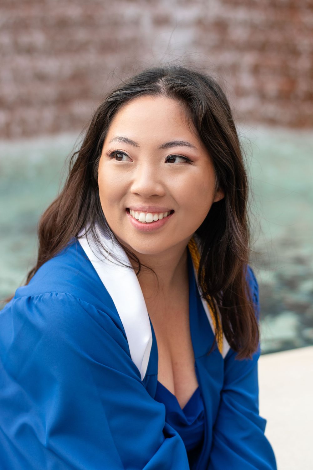Graduate in blue and white academic regalia smiling warmly against a brick wall background.