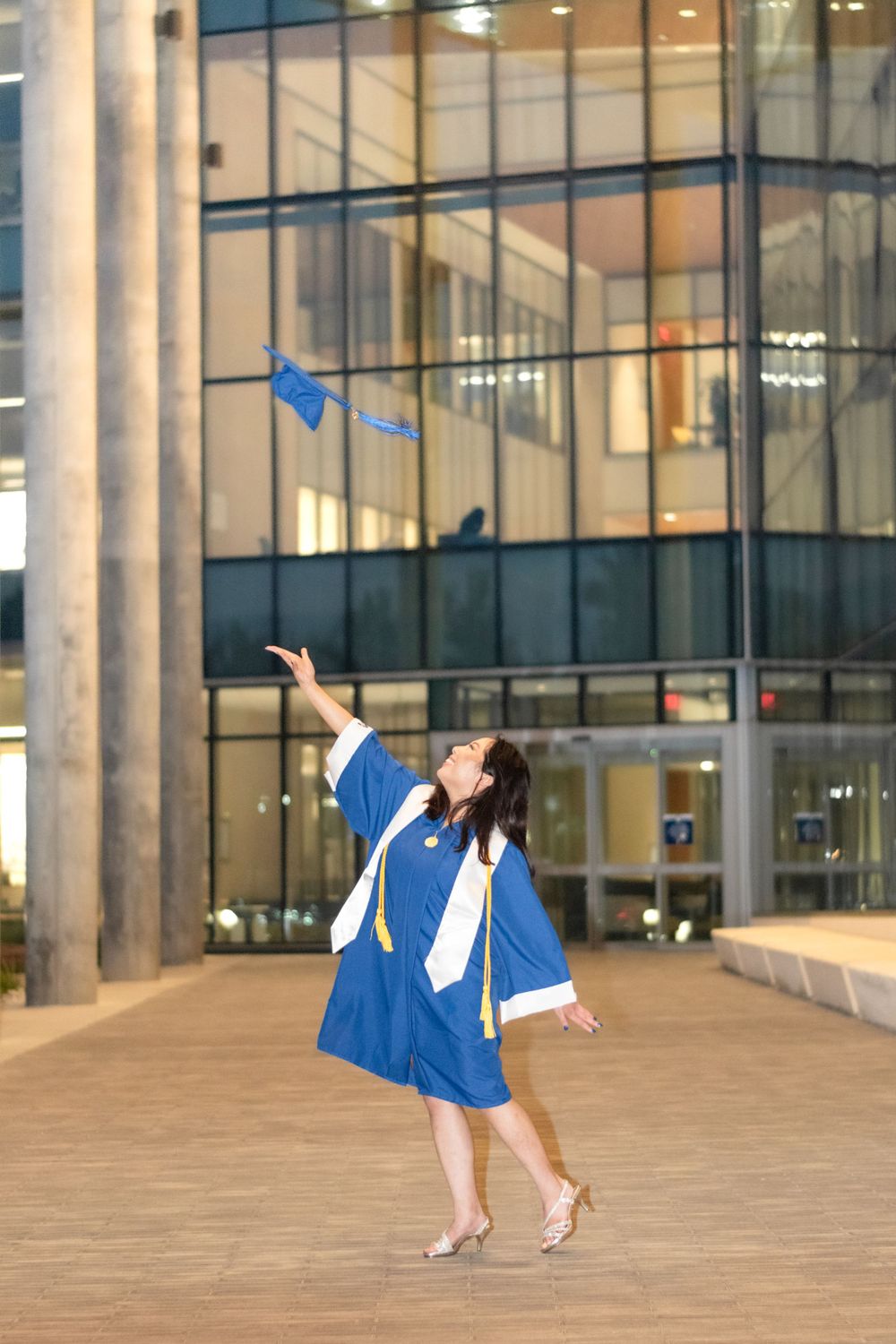 Graduate in blue cap and gown celebrates by tossing graduation cap in front of modern building.