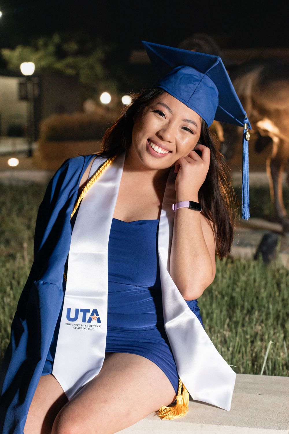 Nighttime graduation portrait with blue cap and UTA sash against illuminated background.