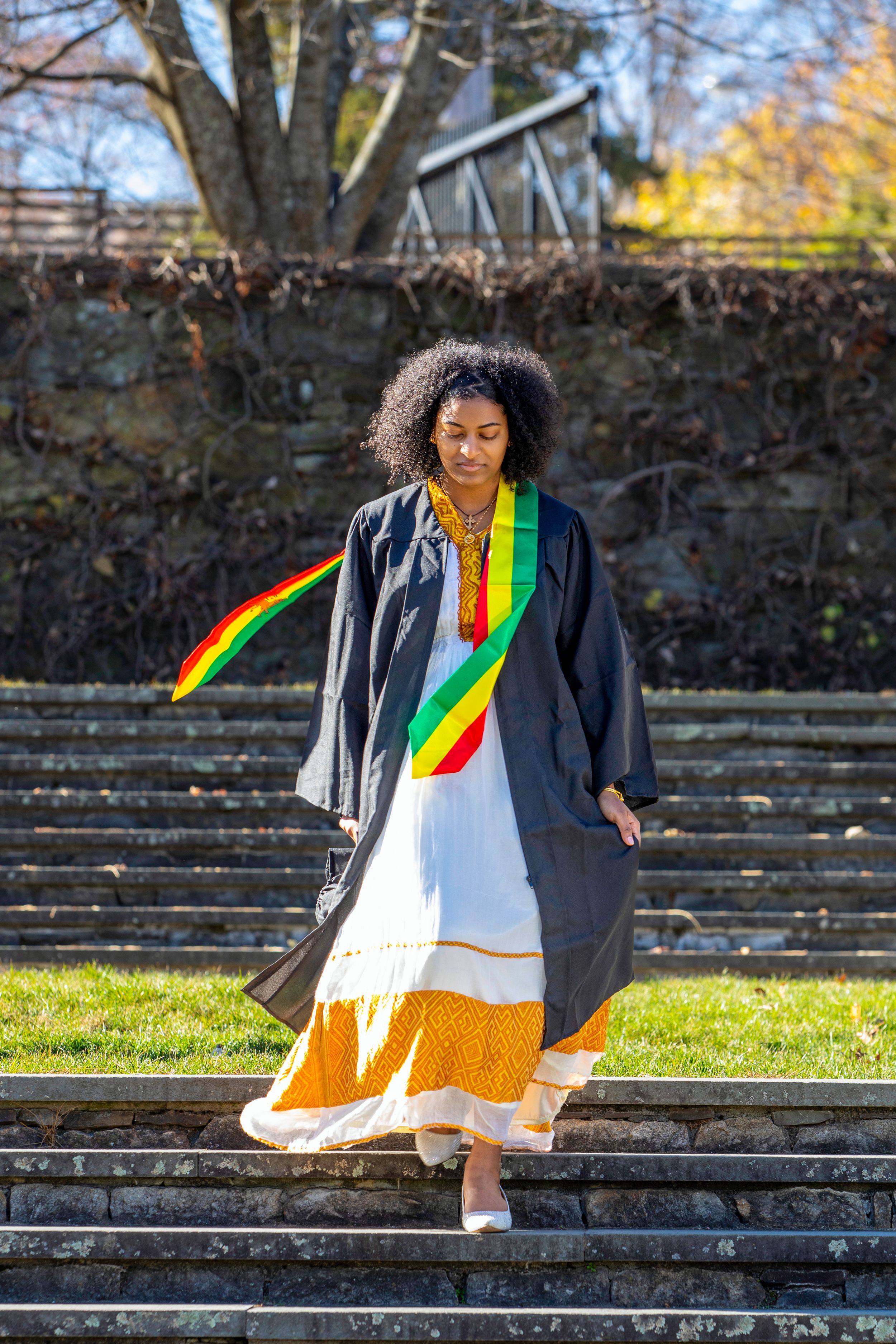 Graduate in traditional dress with colorful sash descending stone steps outdoors.
