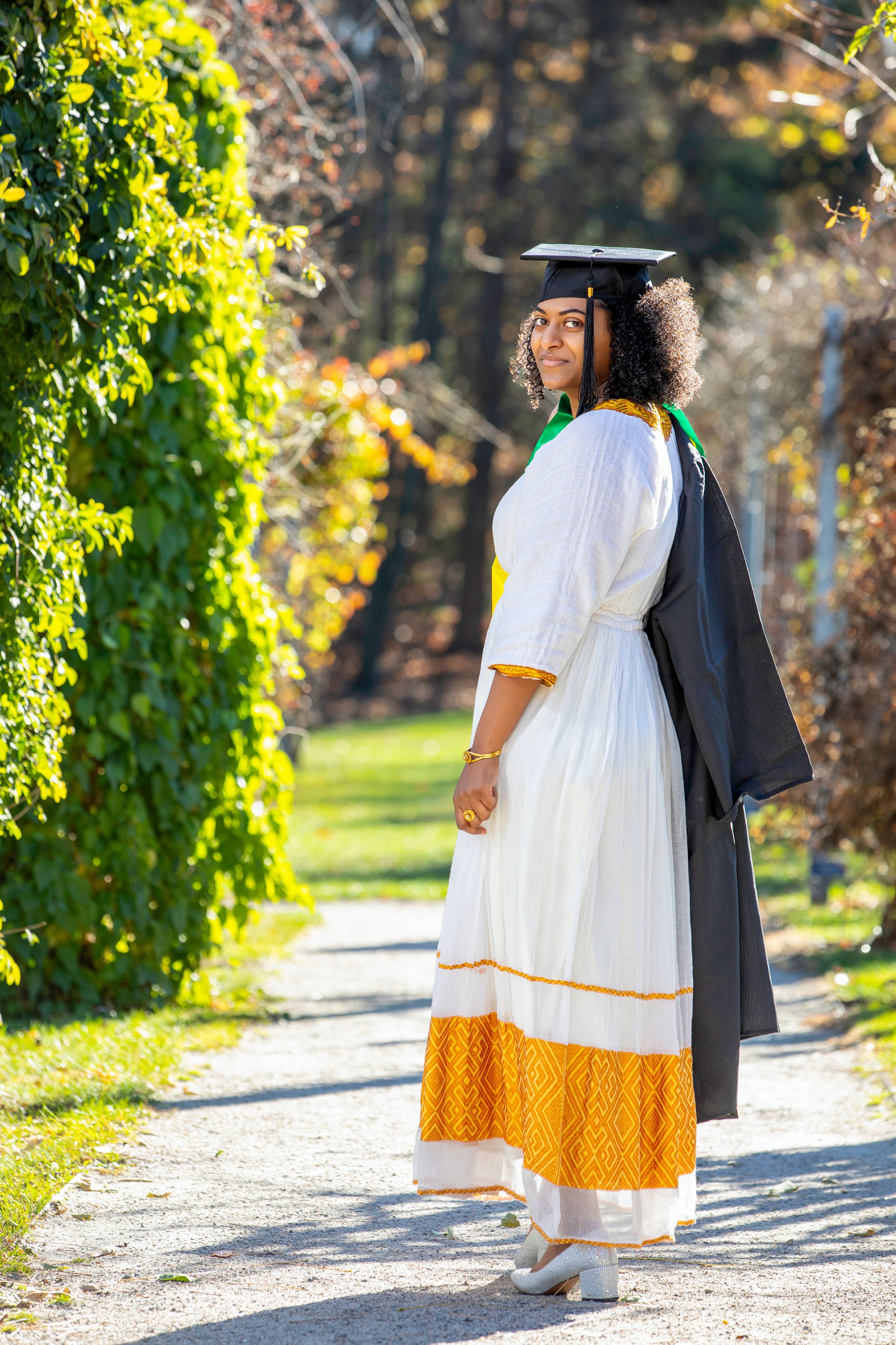 Graduate in academic regalia and traditional white dress posing in garden pathway.