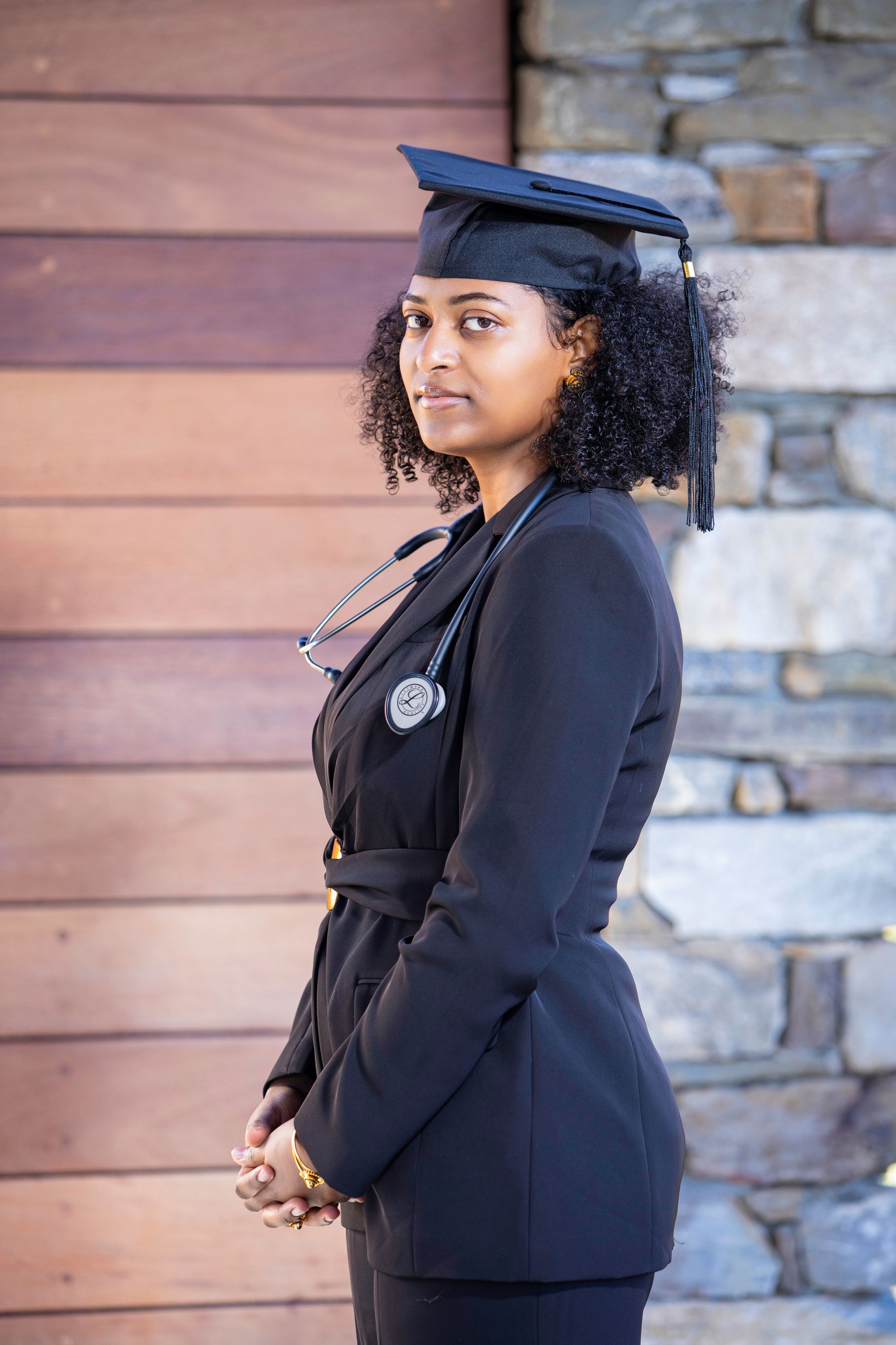 Graduate in black professional attire with stethoscope and graduation cap posing outdoors.