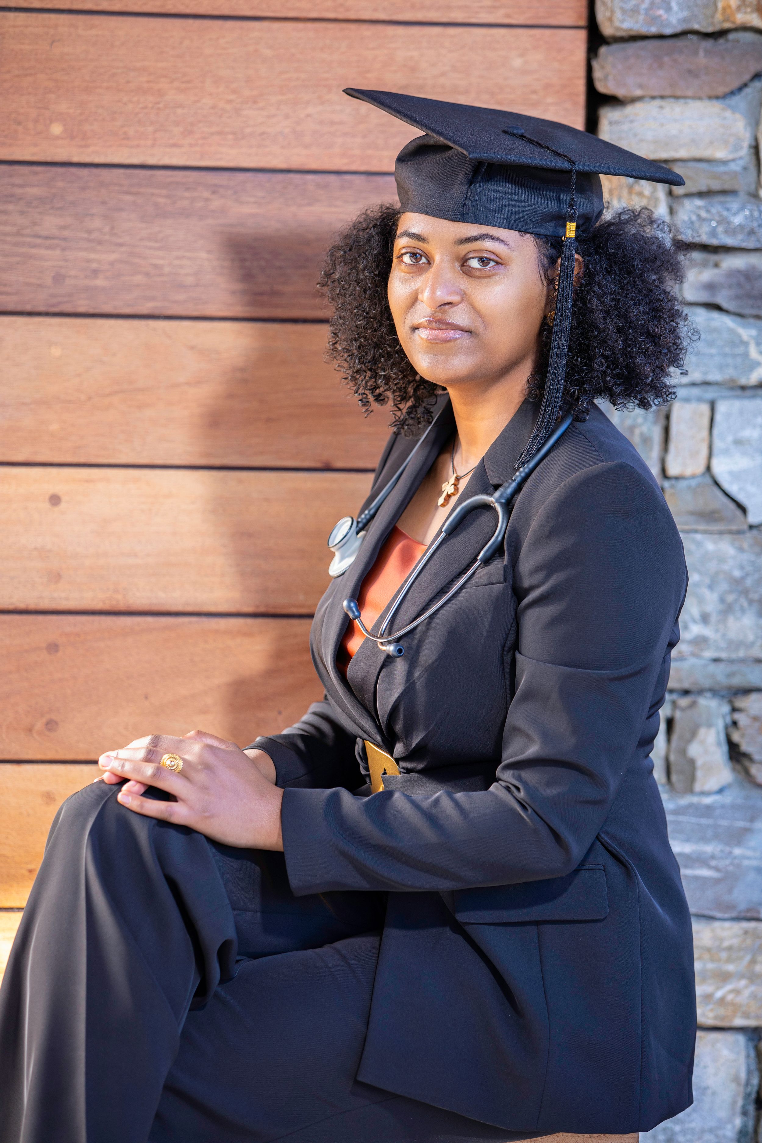 Graduate in formal black attire seated with stethoscope and graduation cap.