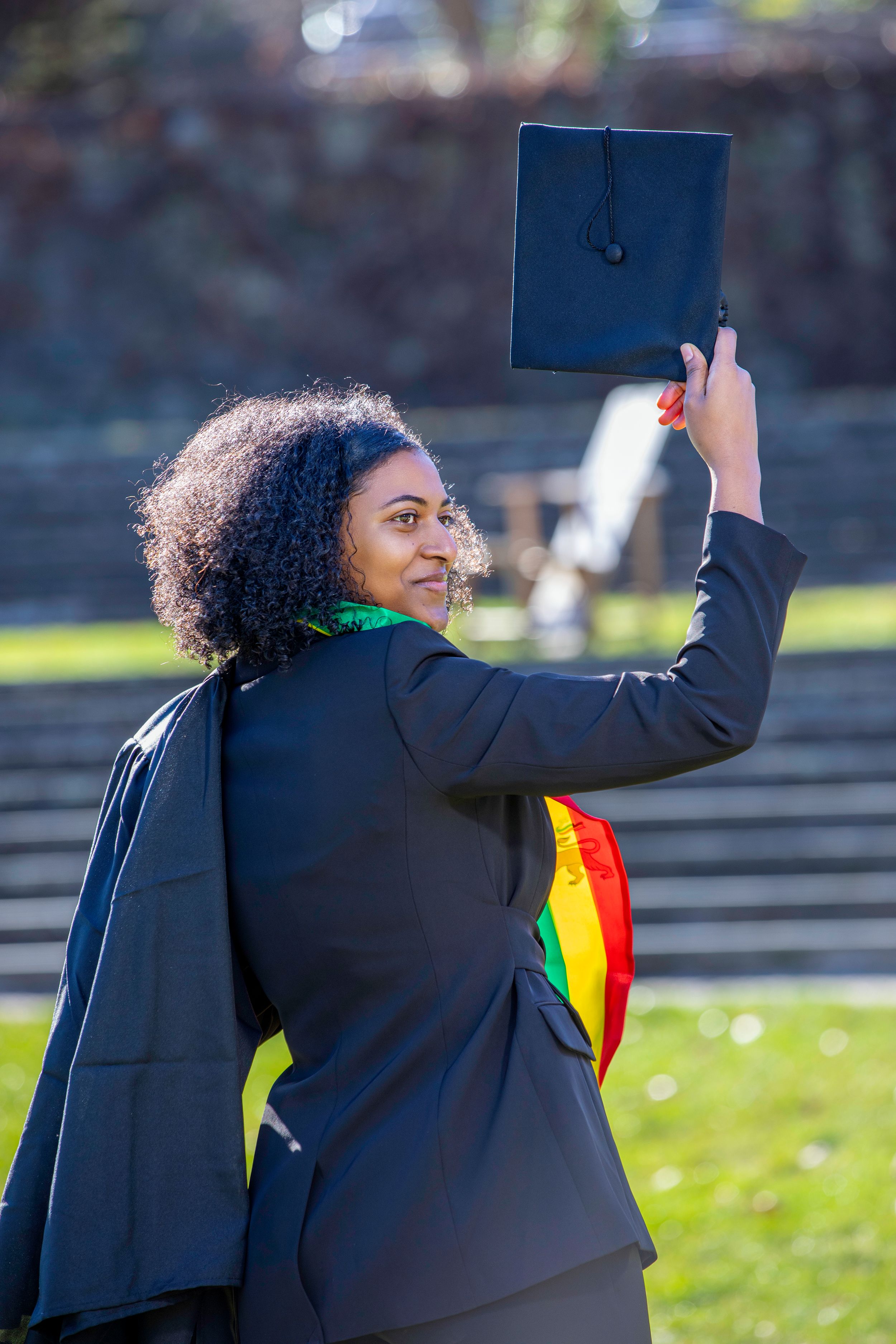 Graduate in academic regalia holding up graduation cap with colorful sash.