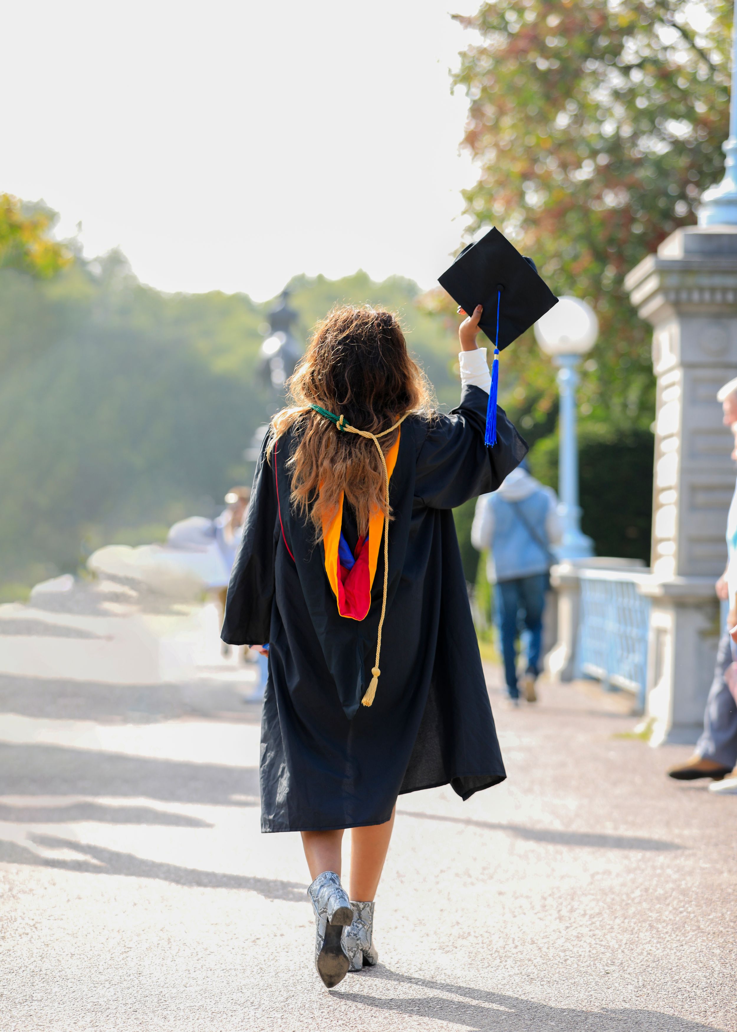 Graduate walking away while holding up graduation cap on campus pathway.