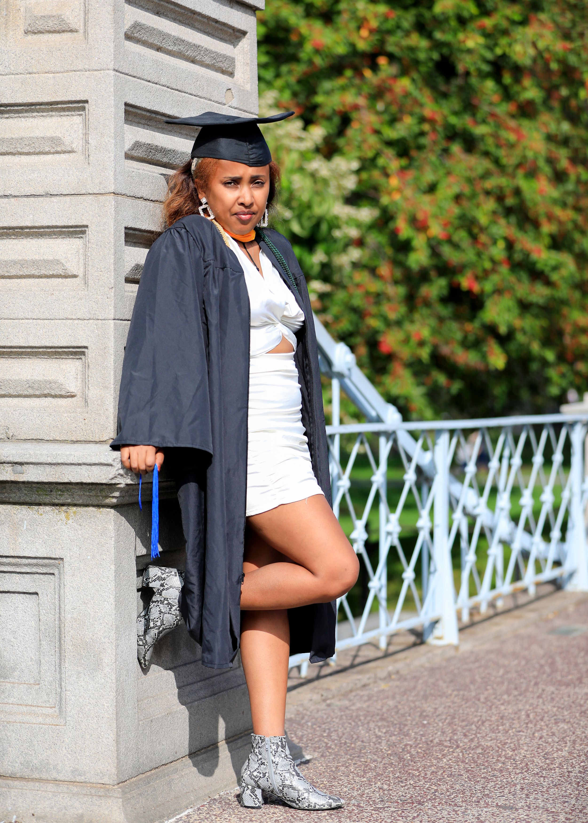 Graduate in cap and gown poses with snake print boots against white column outdoors.
