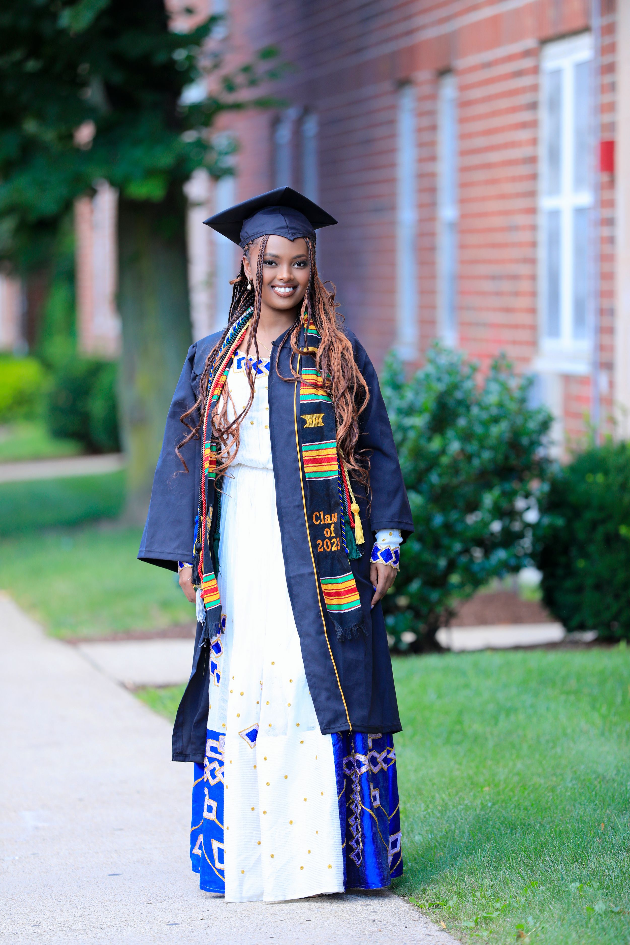 Graduate in decorated cap and gown with colorful kente stole stands gracefully near brick building.