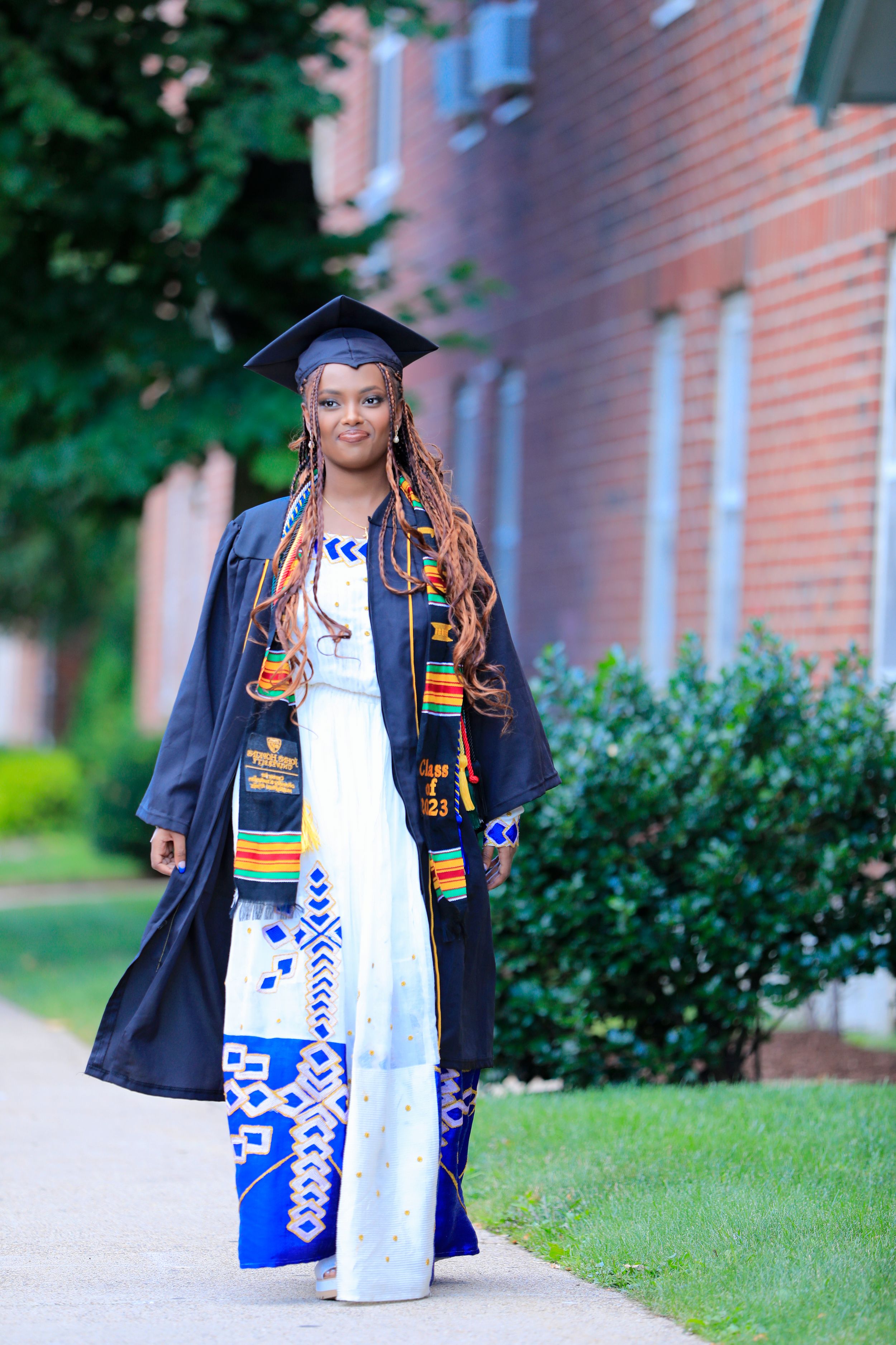 Graduate wearing traditional African-inspired stole and patterned dress stands on campus path.