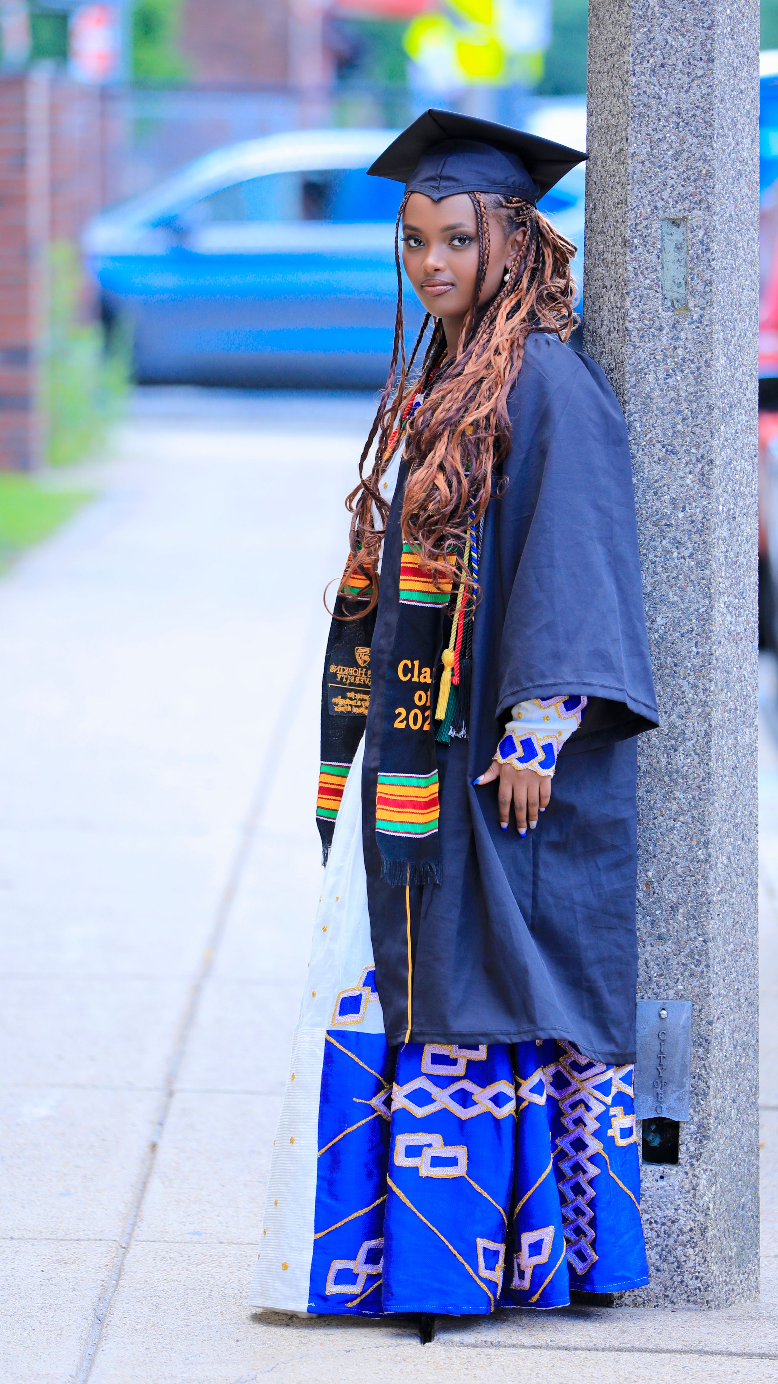 Graduate in traditional academic regalia with kente stole poses against stone column.