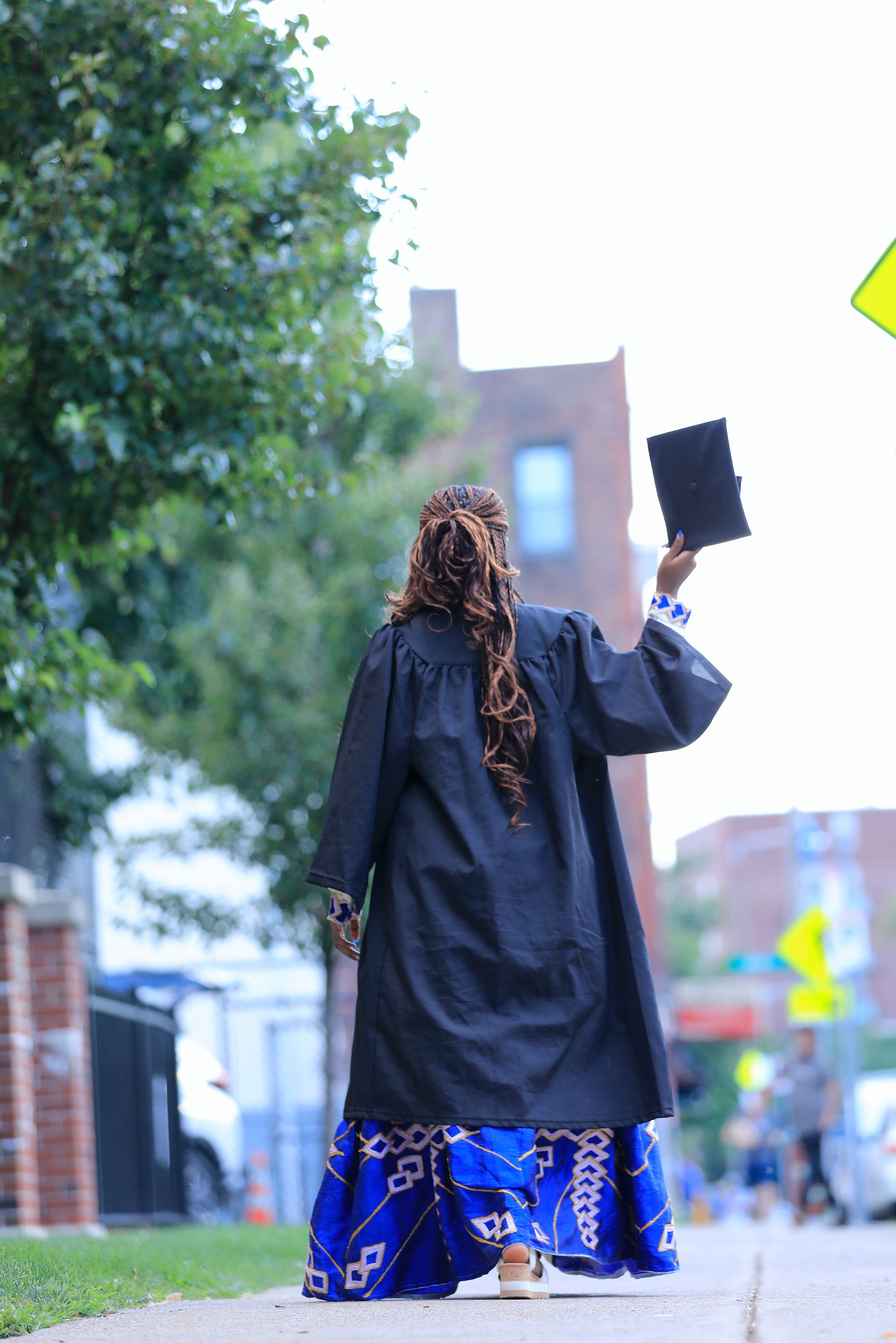 Graduate in ceremonial attire holds diploma high while facing away on campus walkway.