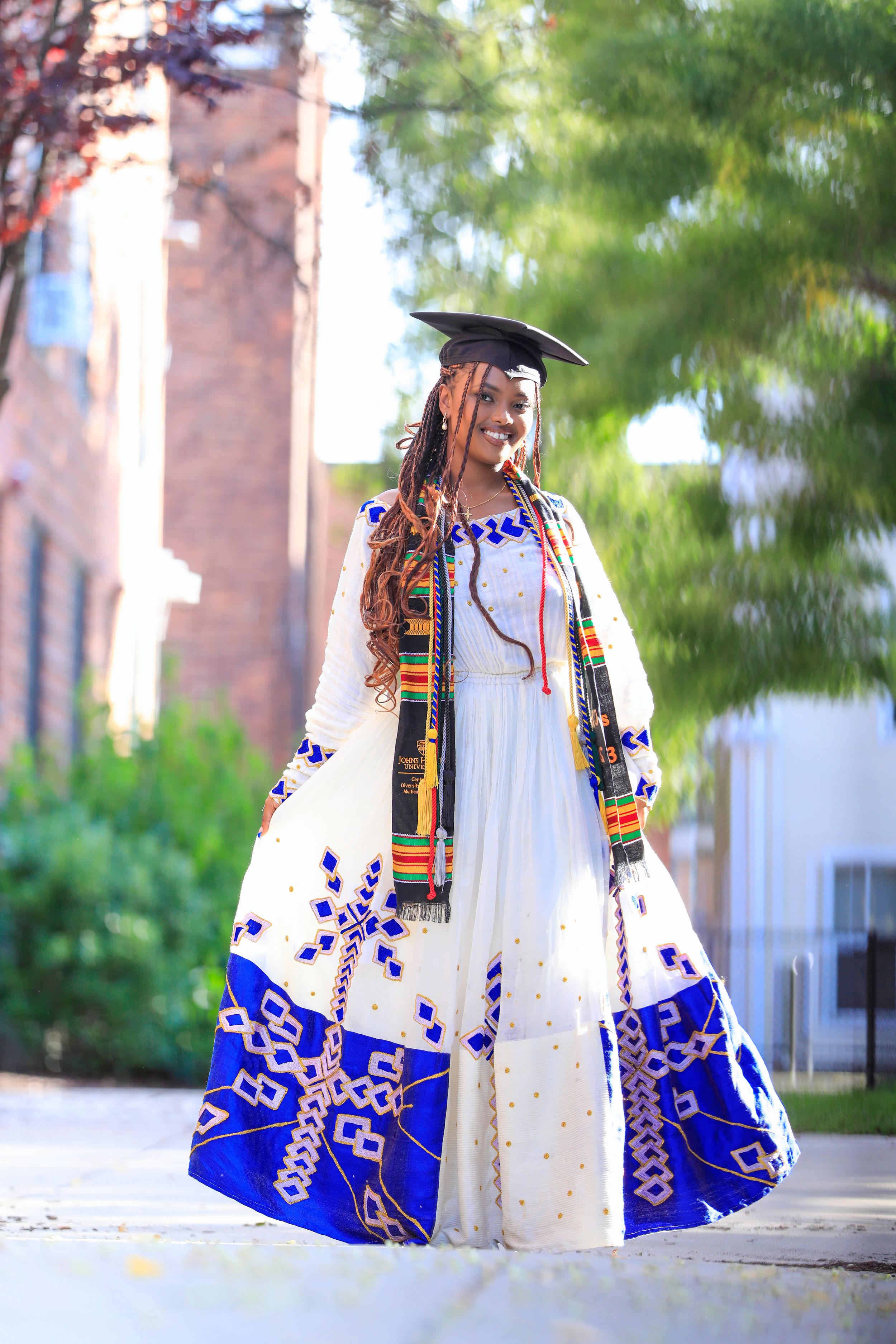Graduate wearing white dress with blue patterns and graduation cap poses outdoors.