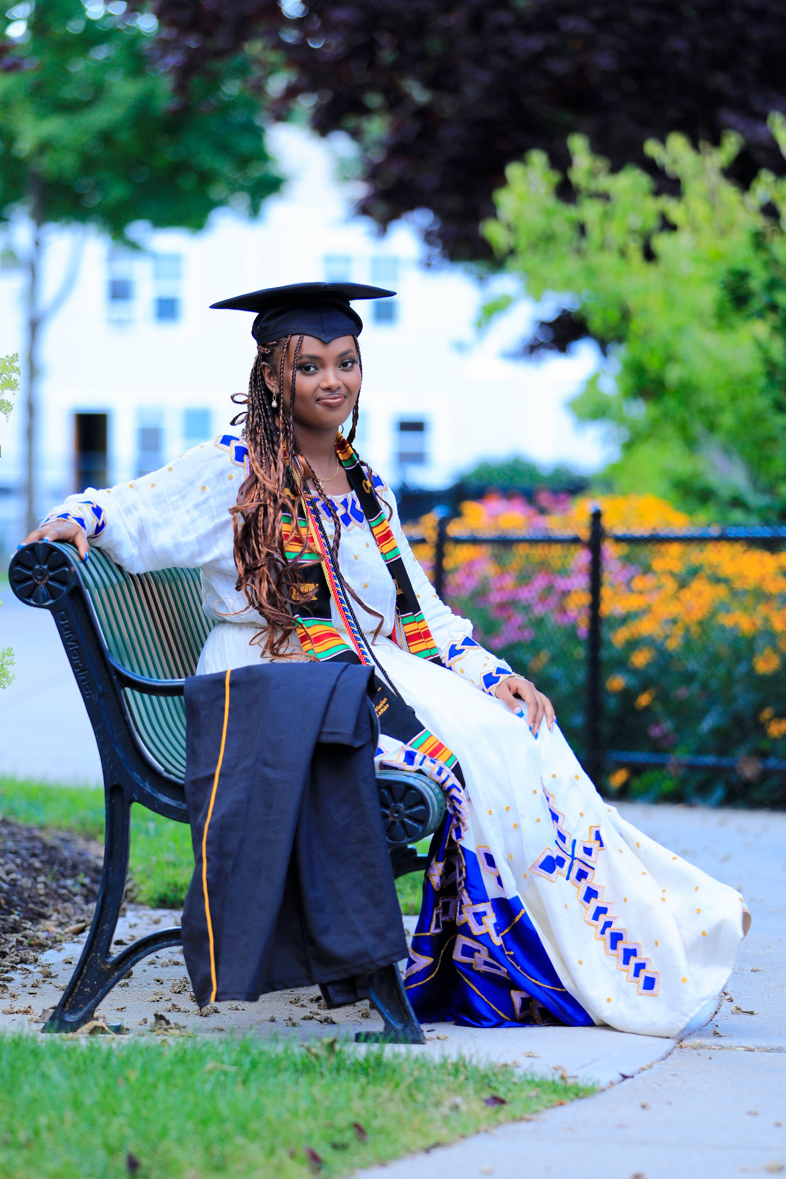 Graduate in decorated regalia sits on park bench surrounded by campus greenery.
