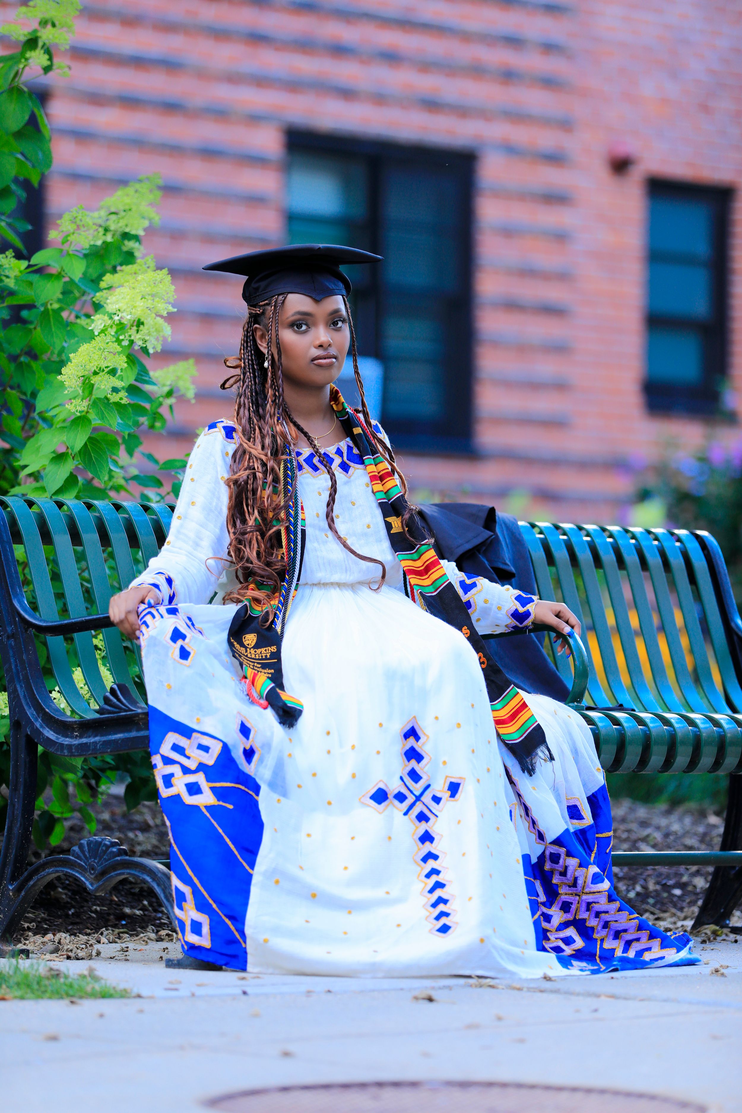 Graduate in white patterned dress and graduation cap sits on campus bench.