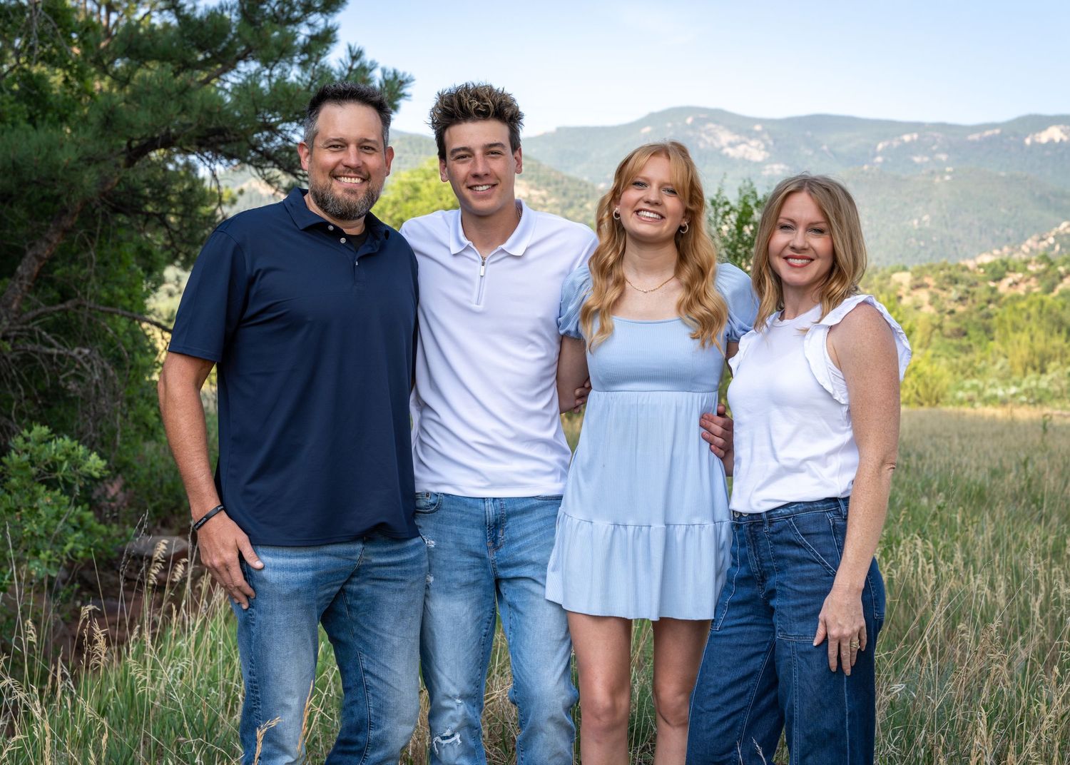 Family portrait of four people standing together outdoors with mountain landscape in background.