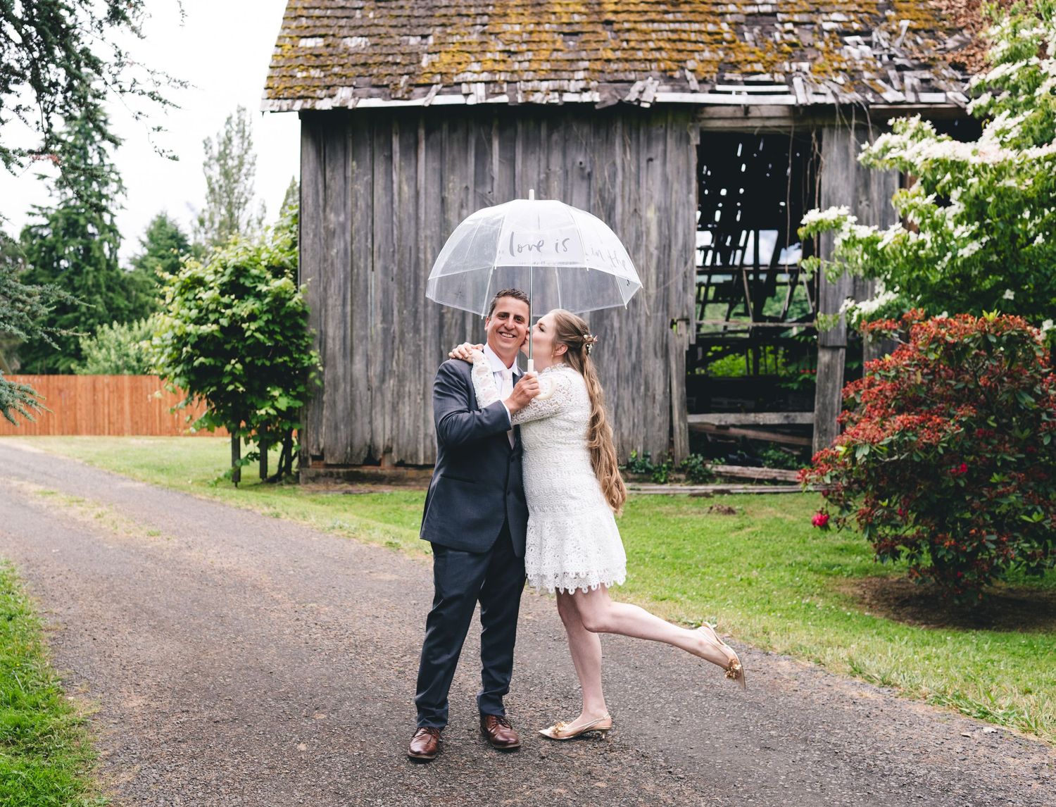 A couple poses with a white umbrella in front of a rustic barn on their rainy wedding day.