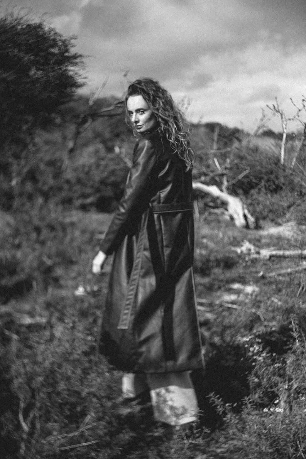 Figure in dark coat walking through windswept outdoor landscape in black and white.