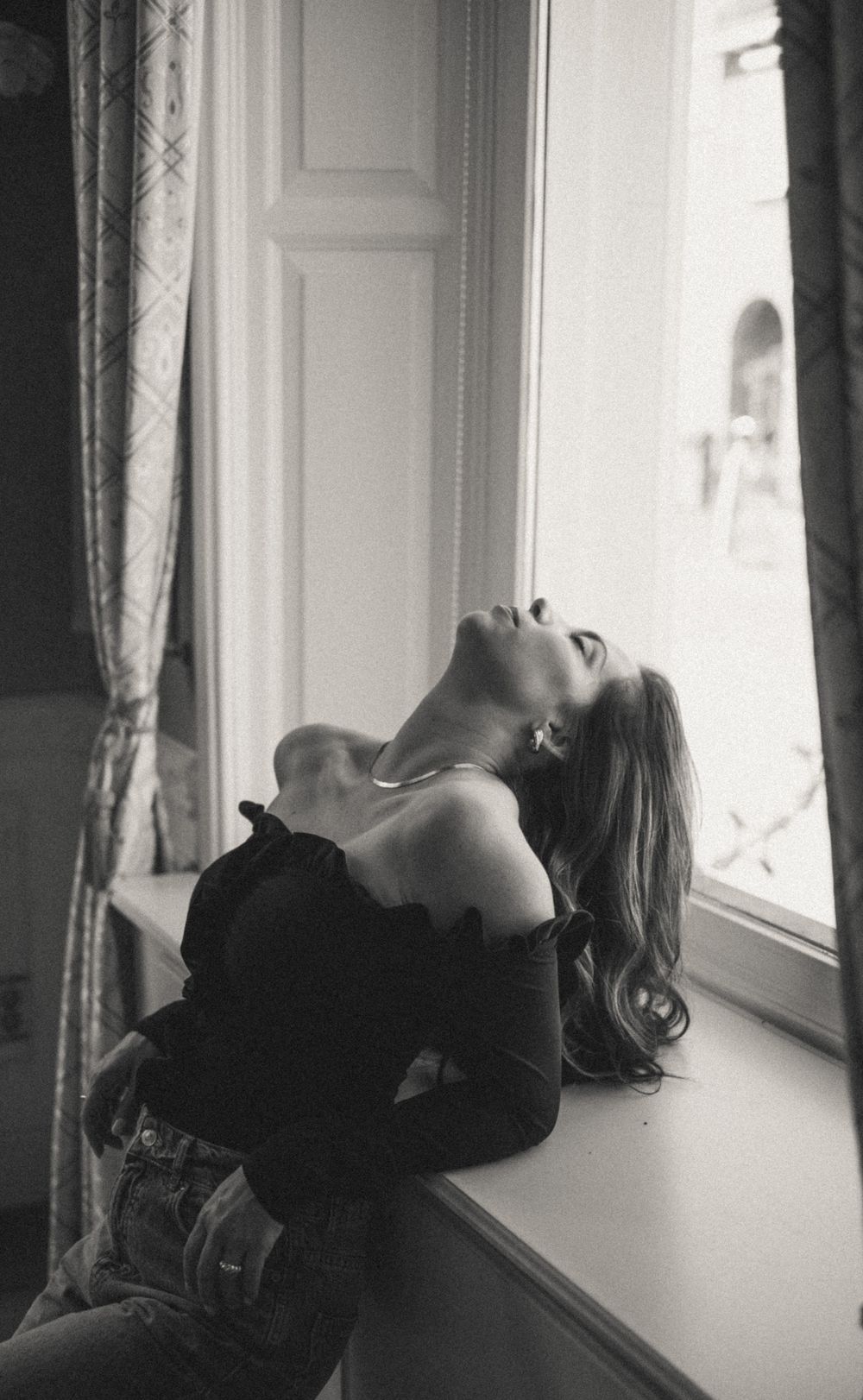 Dramatic black and white photo of figure reclining on windowsill in dark clothing.