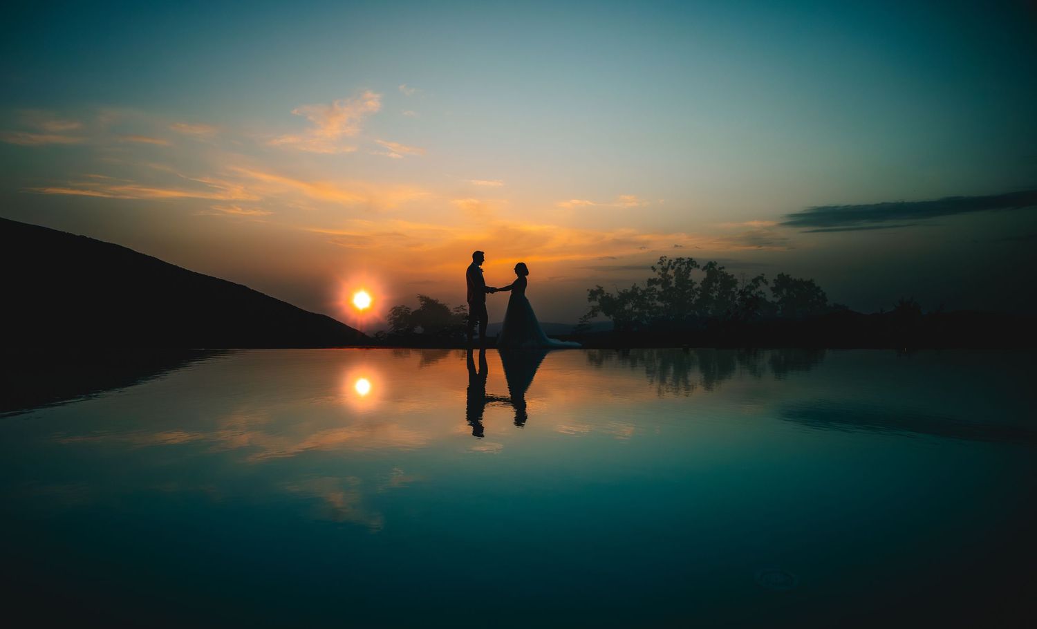 Silhouettes of two people holding hands at sunset reflected in still water creating a mirror-like effect. In Assisi, Italy
