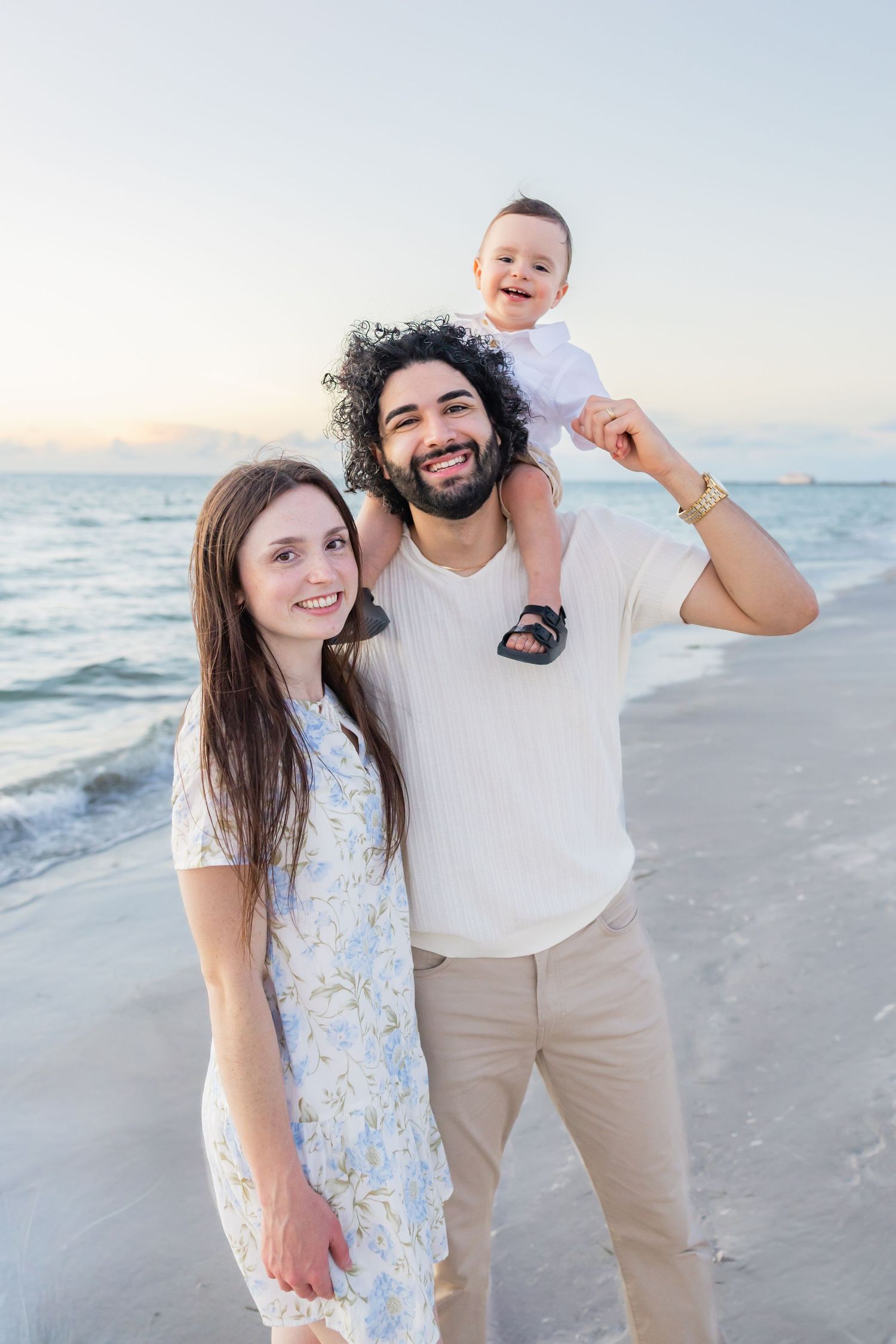 Happy family portrait on beach at sunset, father carrying baby on shoulders beside smiling mother.