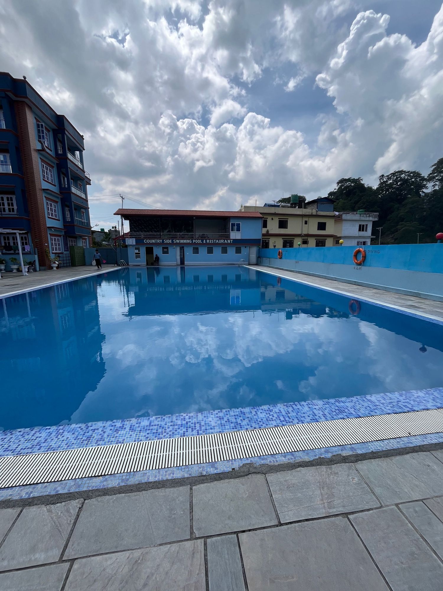 Large swimming pool reflecting cloudy blue sky with modern buildings in background.