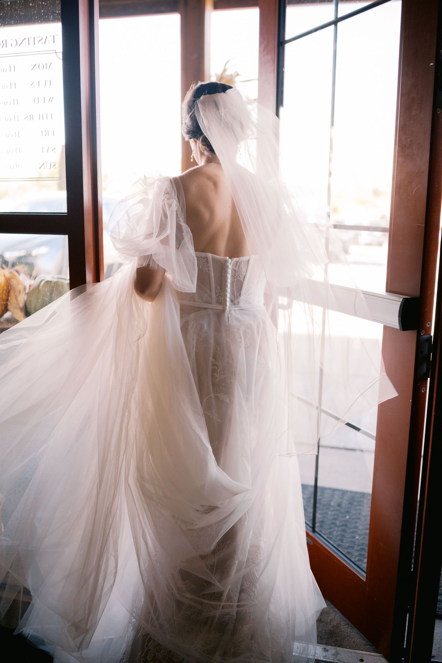 A bride in a flowing white tulle wedding gown with an open back design stands elegantly by glass doors.