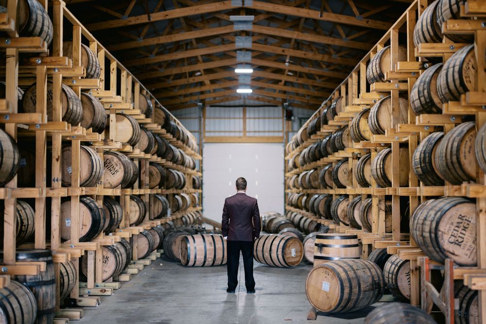A figure stands between rows of wooden whiskey barrels stacked high in a distillery warehouse.