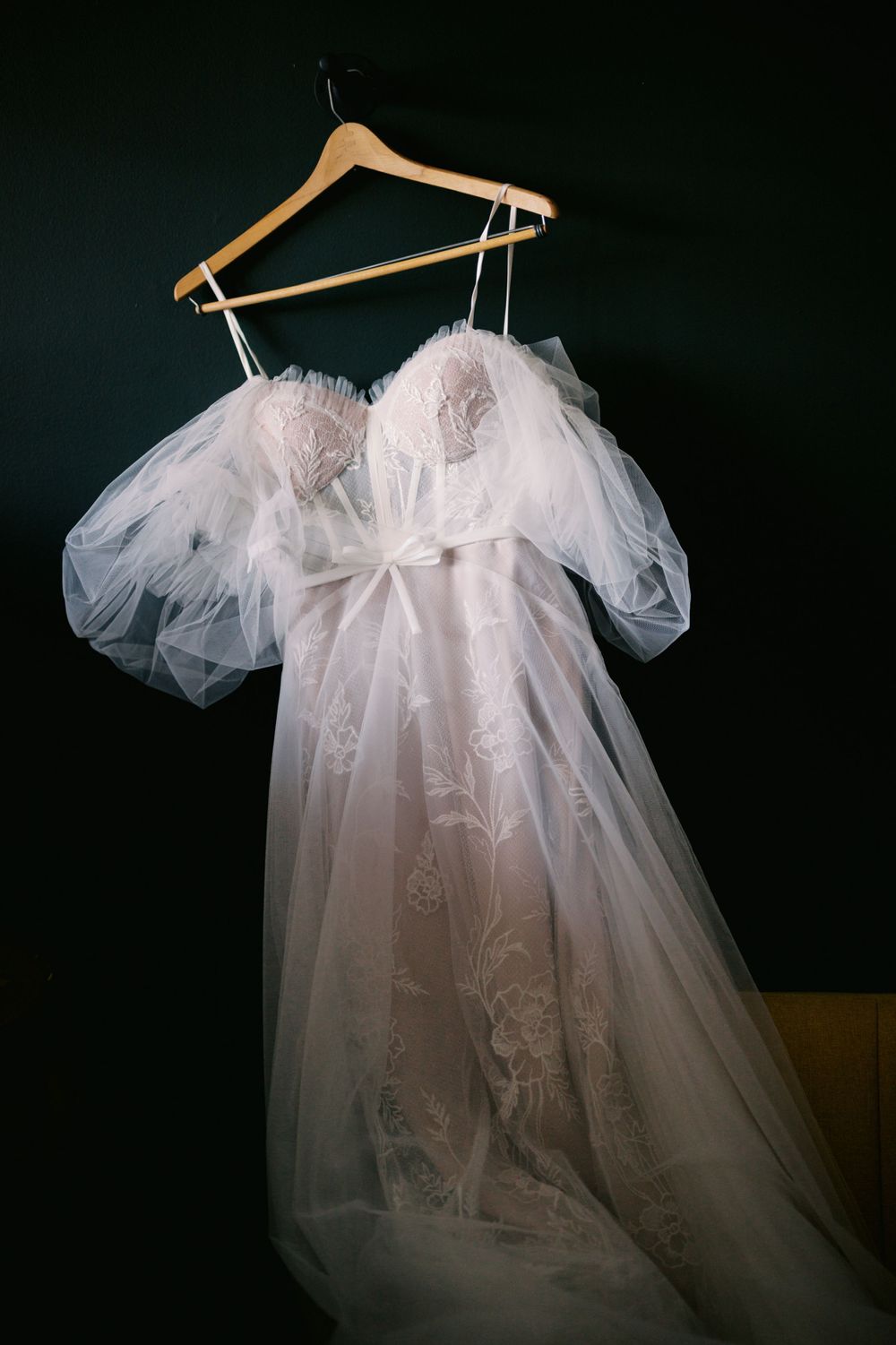 A ethereal white wedding dress with puffy tulle sleeves and lace details hangs on a wooden hanger against a dark background.