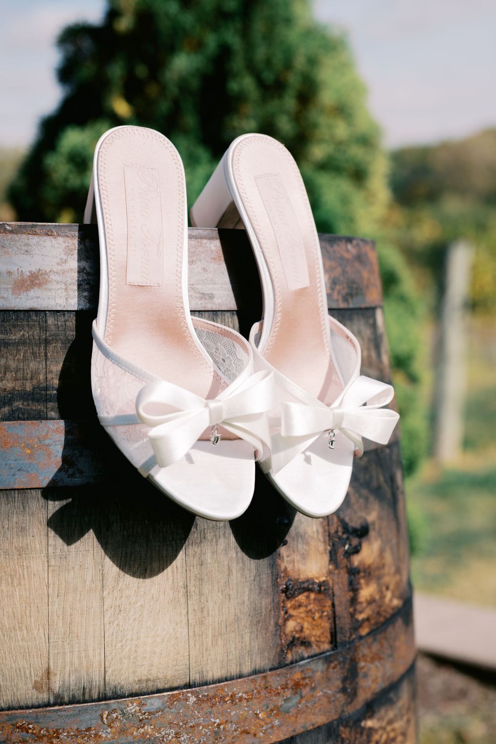 Elegant white bridal heels with bow details rest on a rustic wooden wine barrel in an outdoor vineyard setting.
