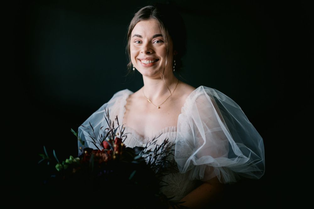A bride in a white puffy-sleeved dress poses with a dark floral bouquet against a moody dark background.