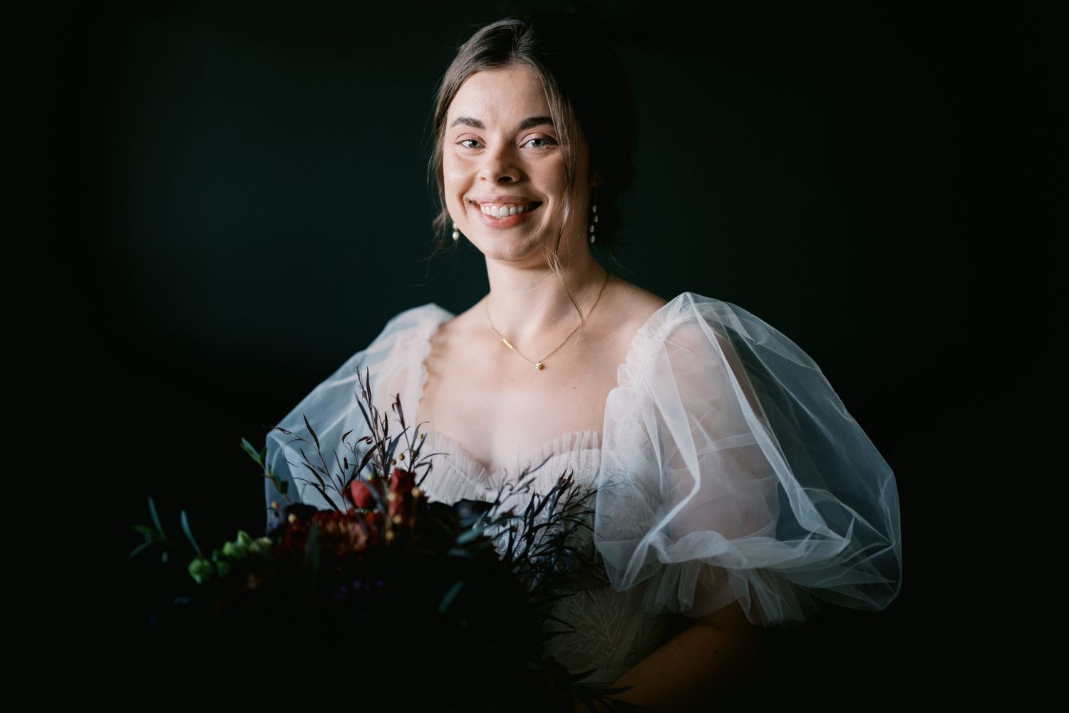 A bride in a white puffy-sleeved dress poses with a dark floral bouquet against a moody dark background.