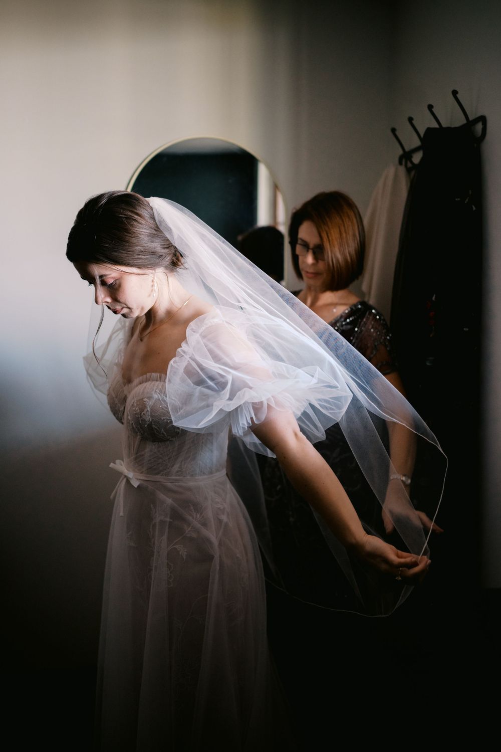 A delicate lace wedding dress and veil are being carefully adjusted in a dimly lit dressing room.