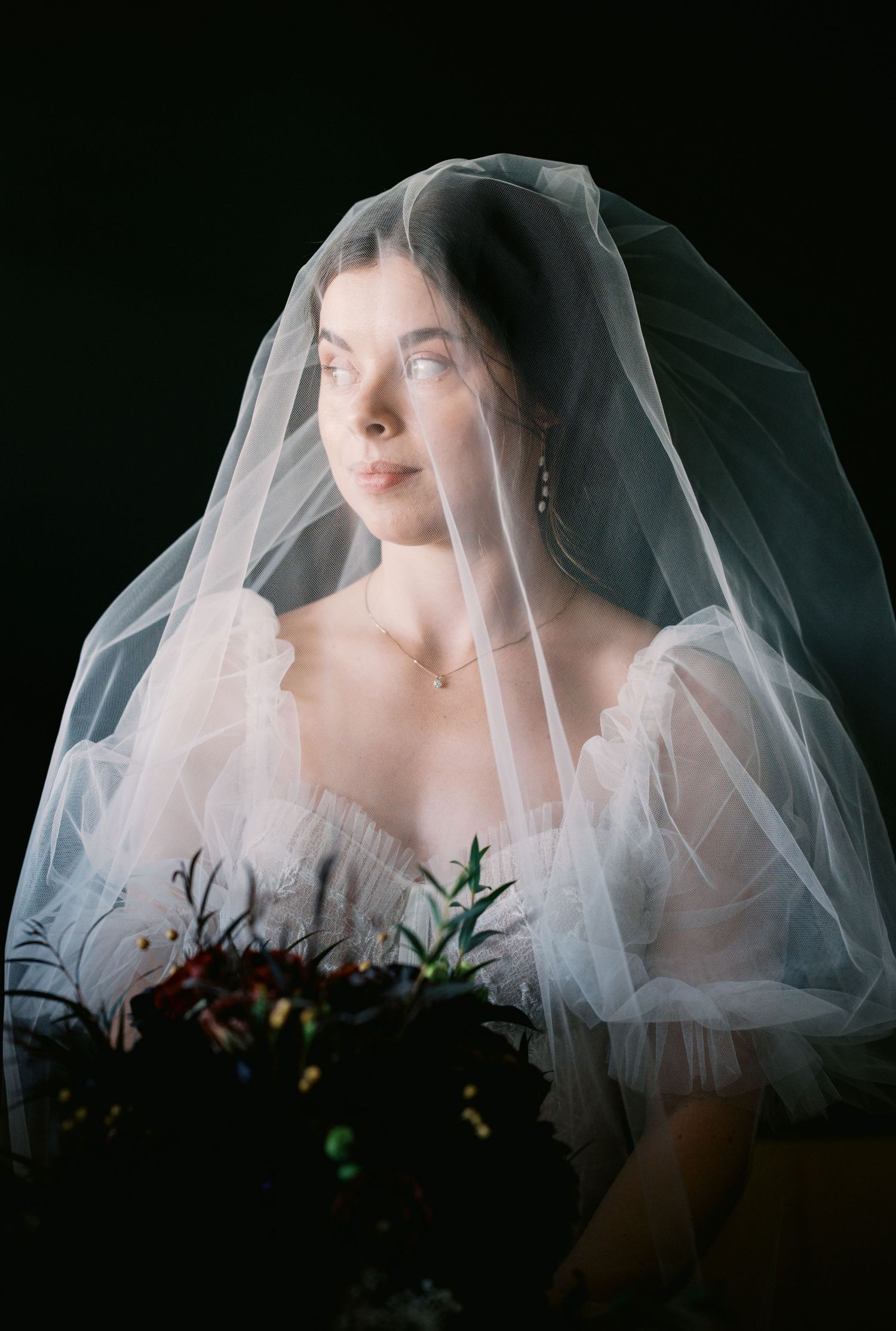Ethereal bridal portrait featuring a sheer white veil and delicate off-shoulder gown against a dark background.