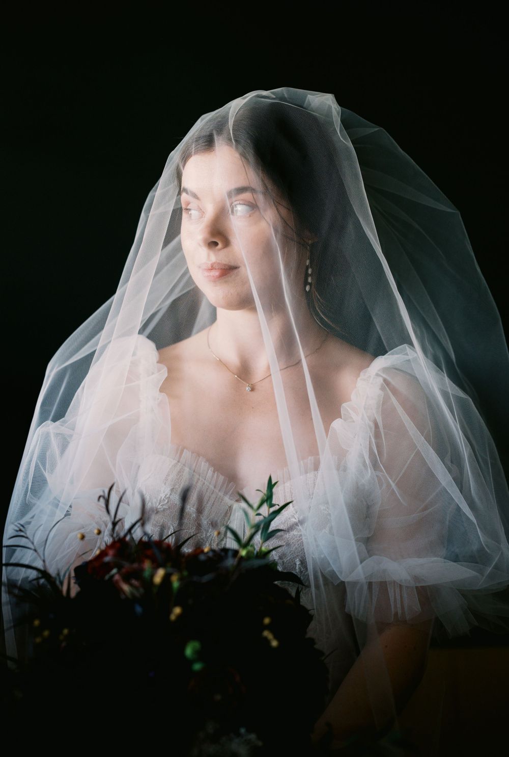 Ethereal bridal portrait featuring a sheer white veil and delicate off-shoulder gown against a dark background.