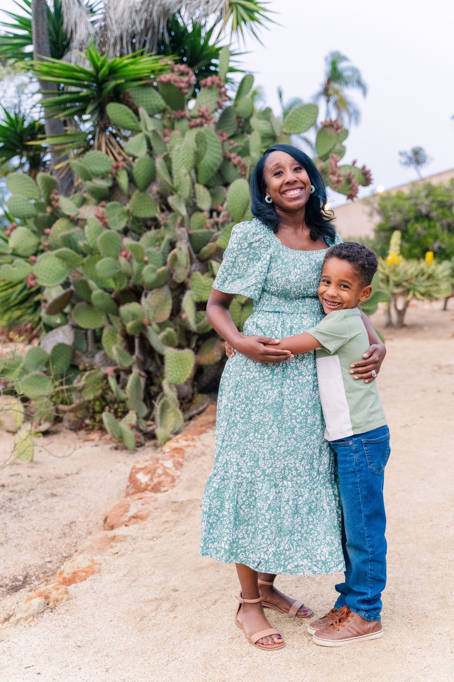 Standing by a cactus on a desert path, a pair share a sweet embrace while wearing casual spring outfits.