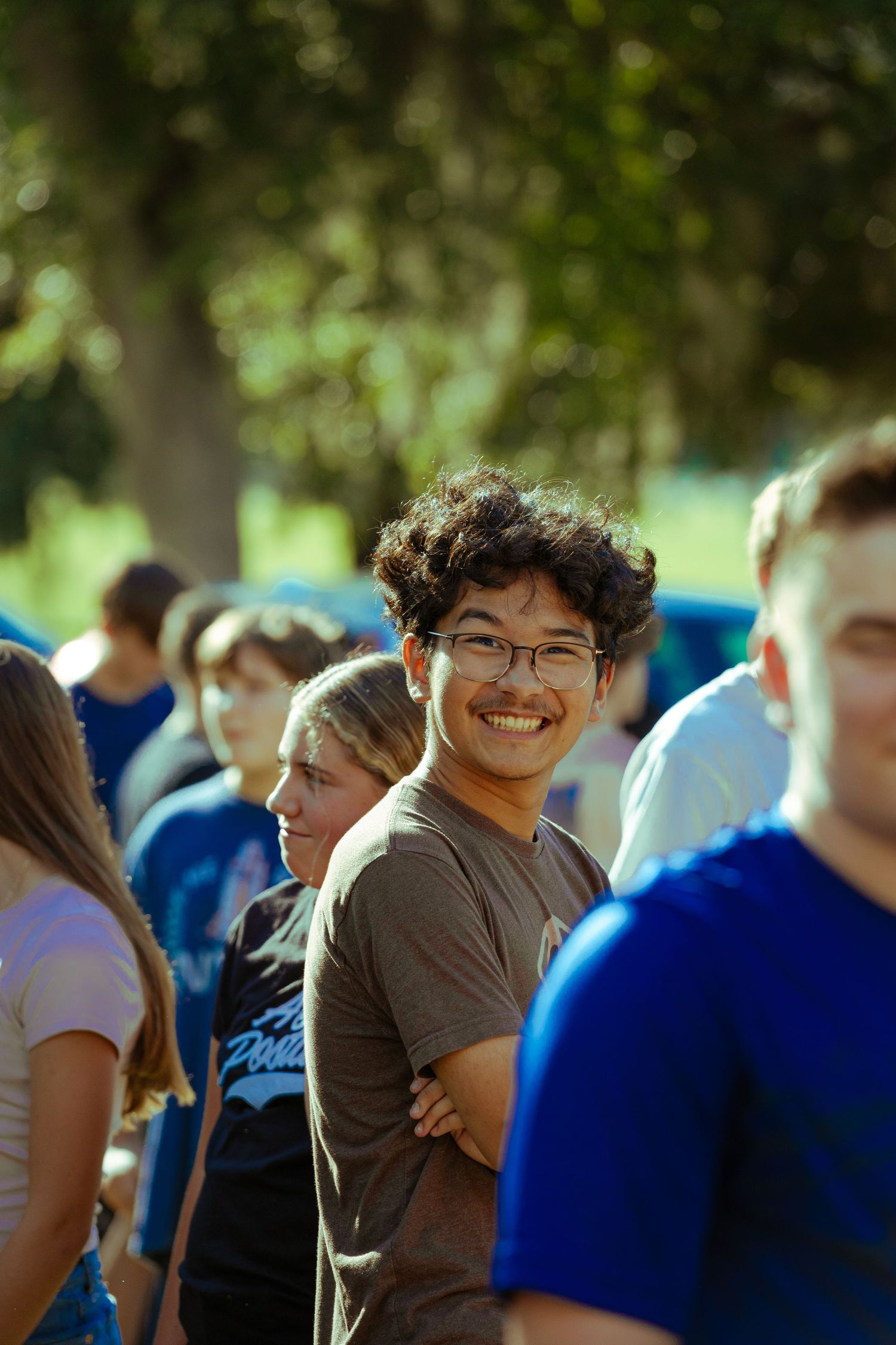 A smiling young man with glasses and curly hair stands with arms crossed among a crowd outdoors.
