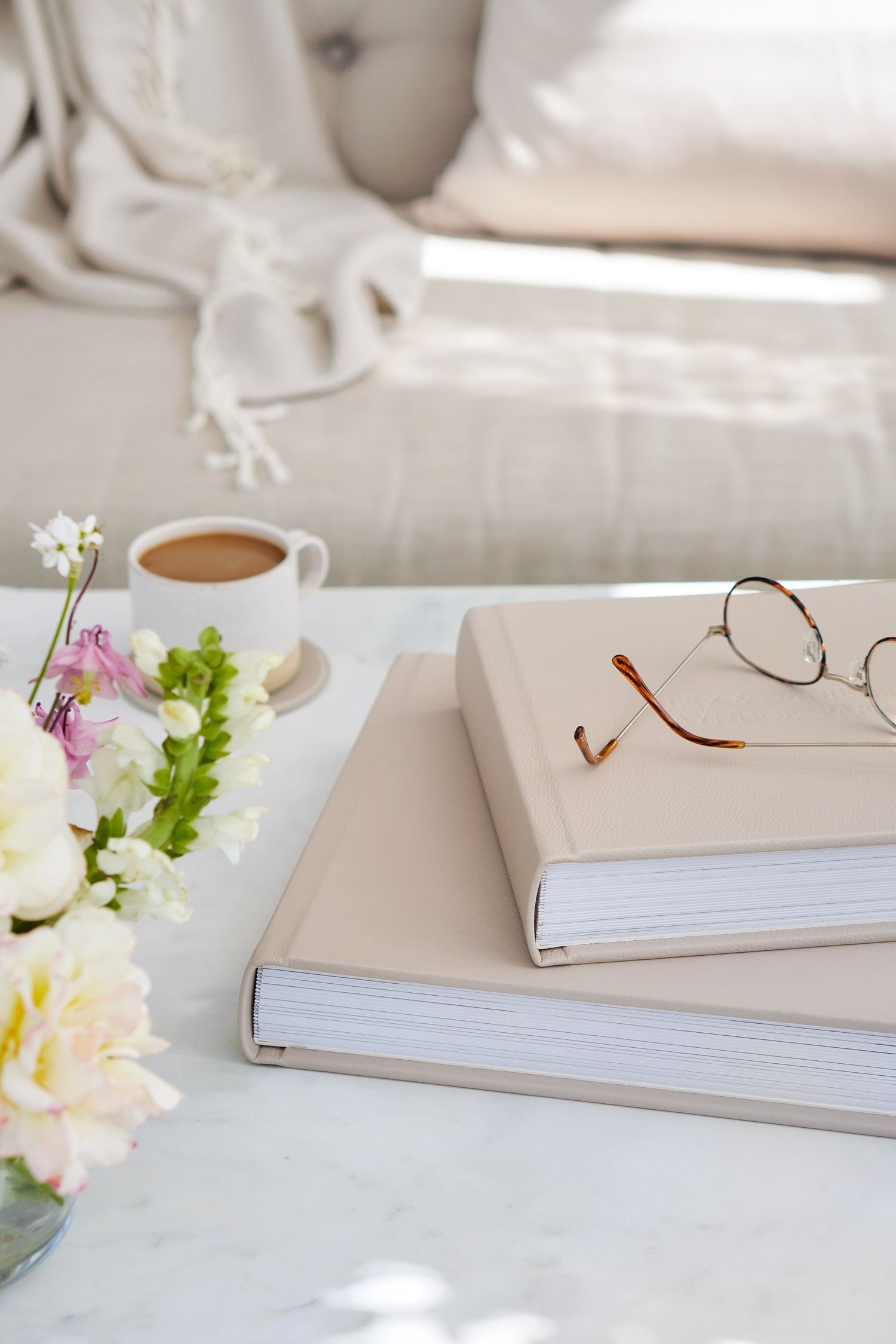 A cozy reading setup with stacked white books, a cup of coffee, and delicate flowers in a vase on a bright table.