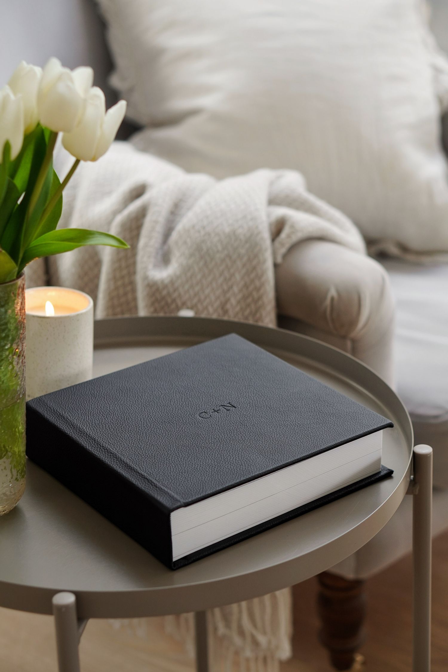 A black leather journal sits on a round table with white tulips and a candle creating a cozy atmosphere.
