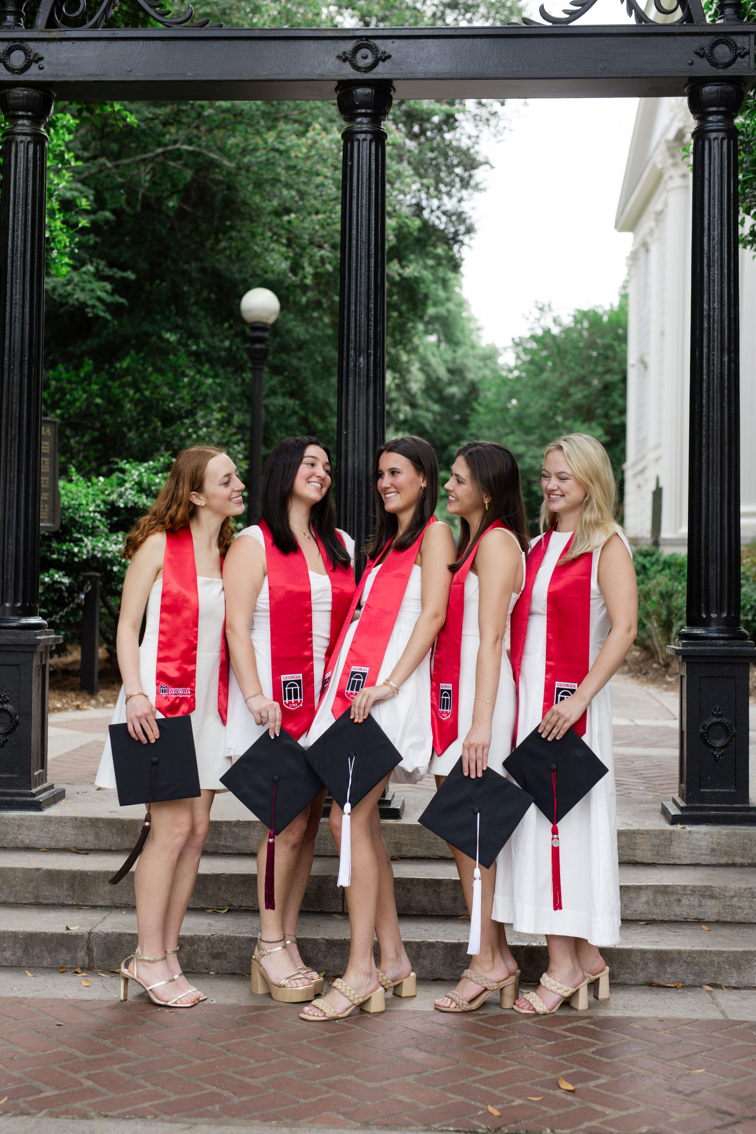 Group of graduates in white dresses and red sashes sit together on brick steps.