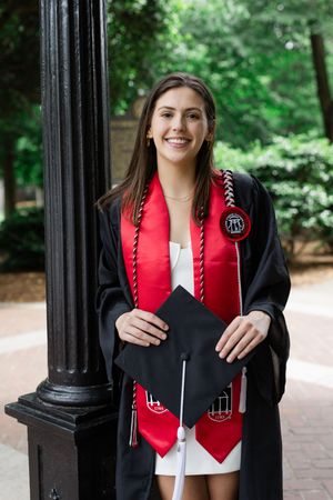 Graduate wearing academic regalia with red stole and graduation cords poses by column.