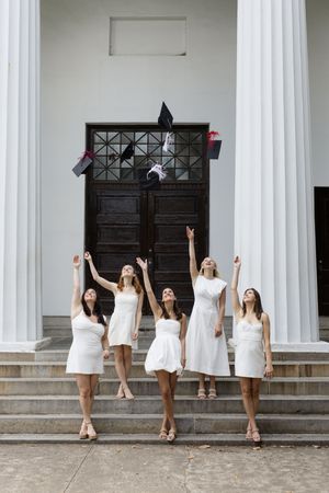 Graduates in white dresses throw their caps in celebration on building steps.