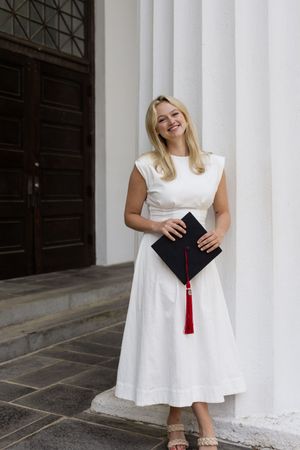Graduate in a white dress poses with a graduation cap and red tassel against white columns.