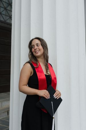 Graduate in black dress and red stole smiles while posing against white columns during graduation photo session.